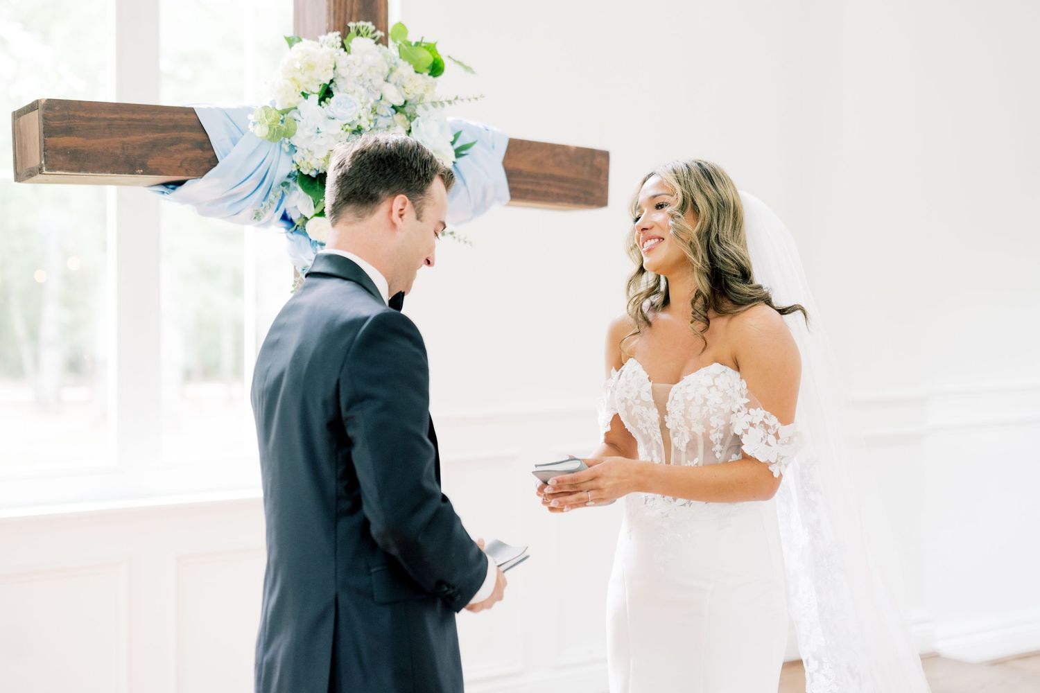 Bride in lace wedding gown exchanging vows with groom beneath floral cross in bright white chapel.