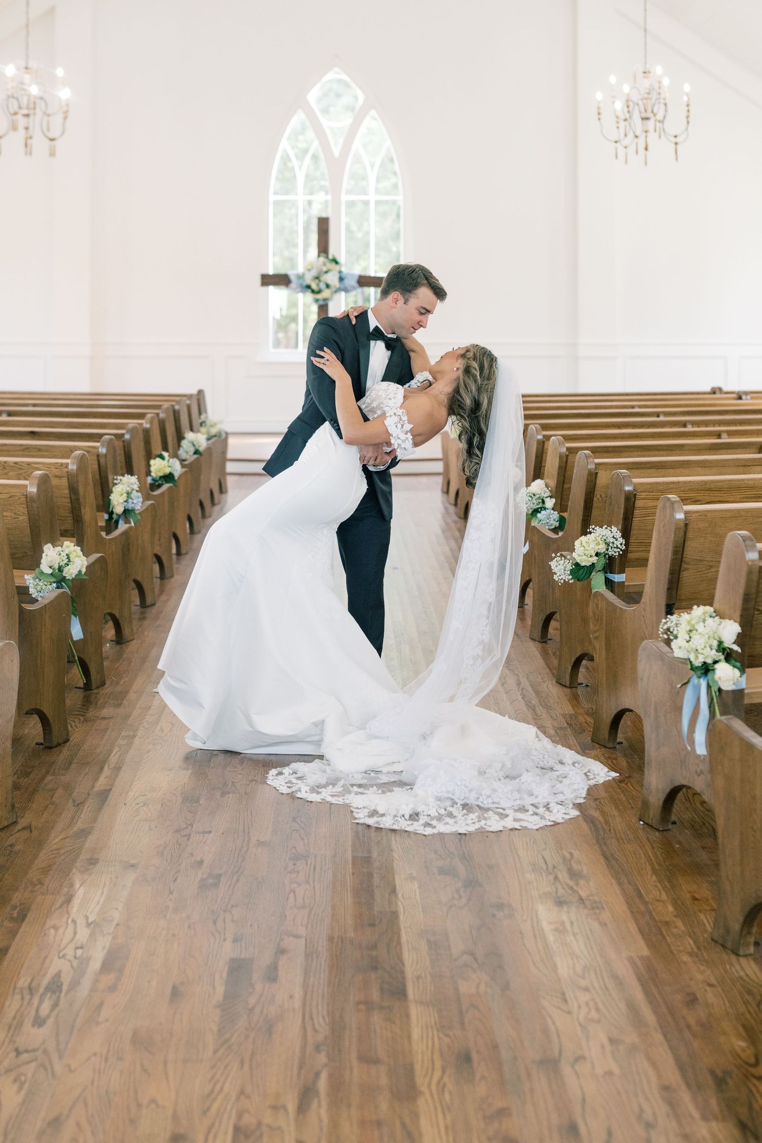 Groom dips bride in white gown and veil in elegant white chapel aisle.