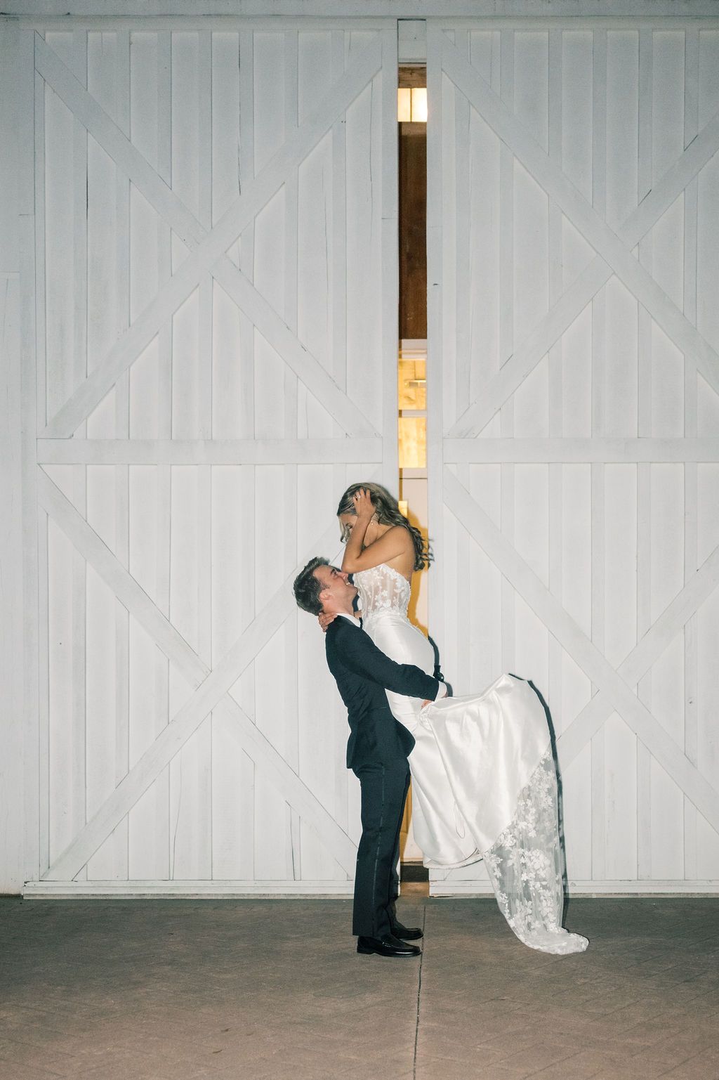 Groom lifting bride in white wedding gown before large white barn doors in a romantic wedding photo.