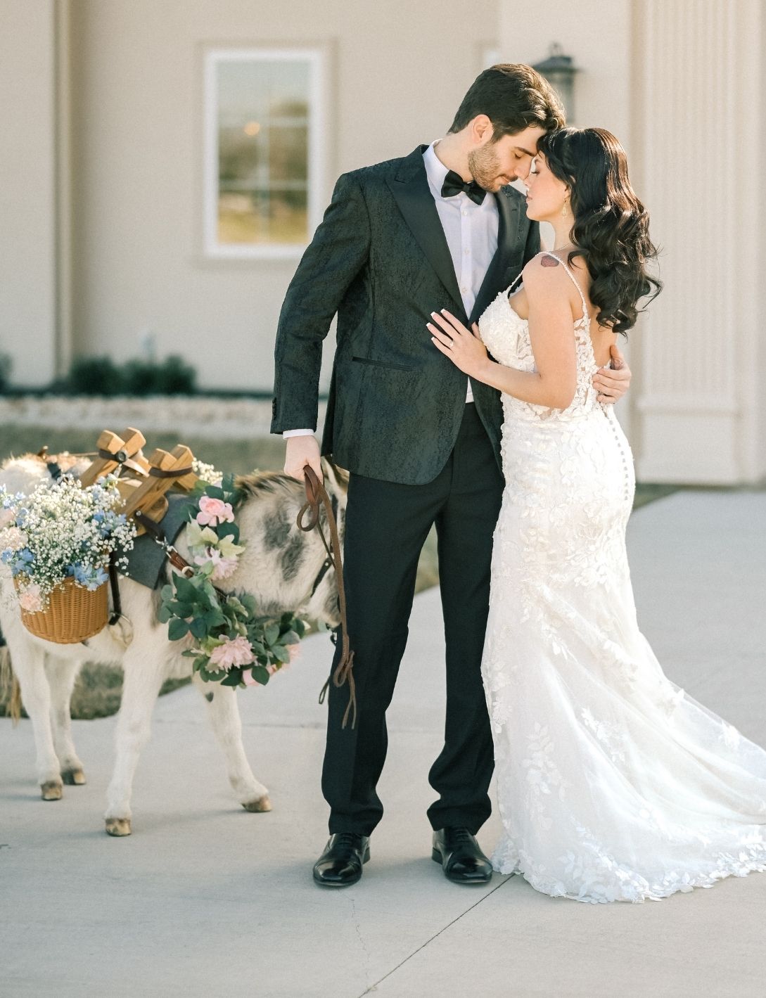 Bride and groom share romantic moment beside a flower-adorned donkey at elegant outdoor wedding venue.