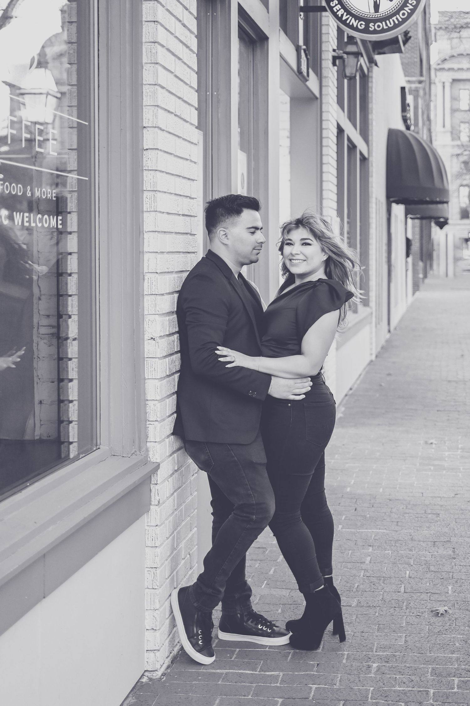 Black and white photo of a stylish couple embracing against a brick building storefront.