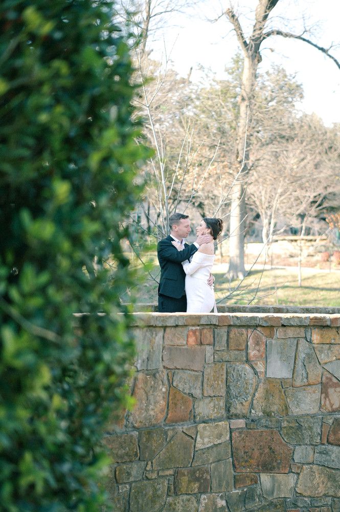 Bride and groom embracing and kissing atop a stone wall surrounded by trees in romantic outdoor setting.