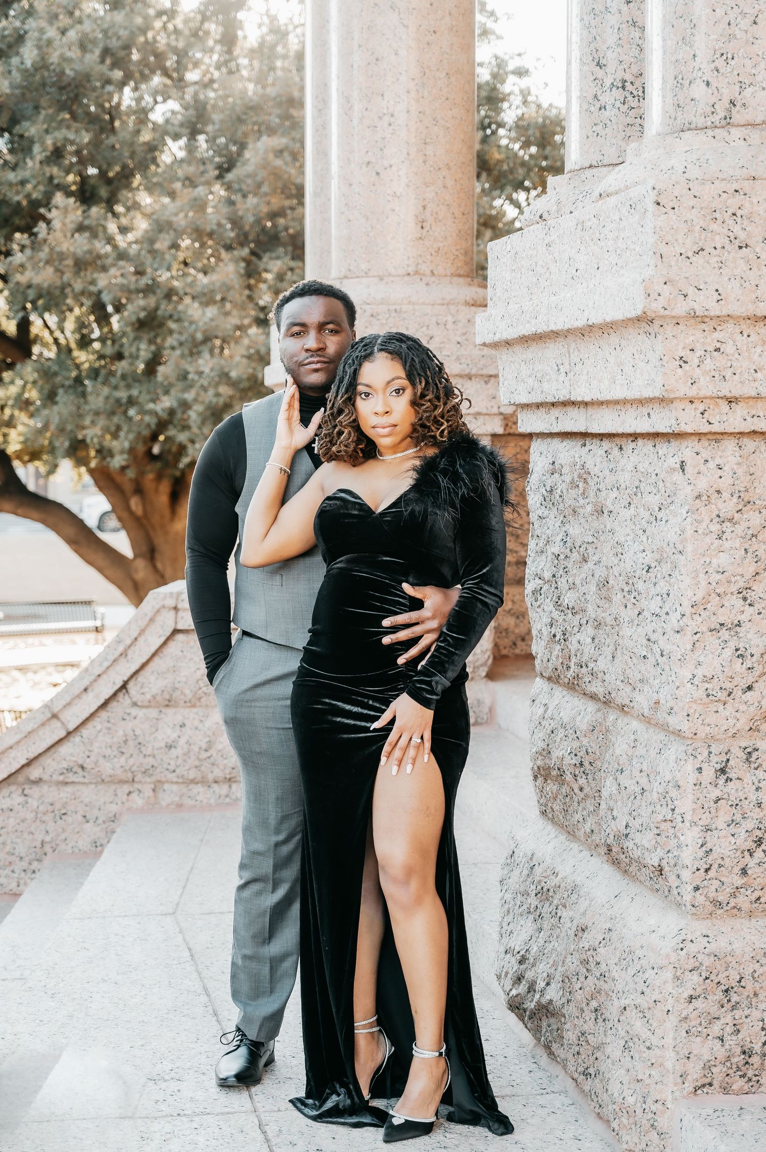 Elegant couple posing by stone columns, man in gray suit and woman in black velvet slit gown with feather sleeve.