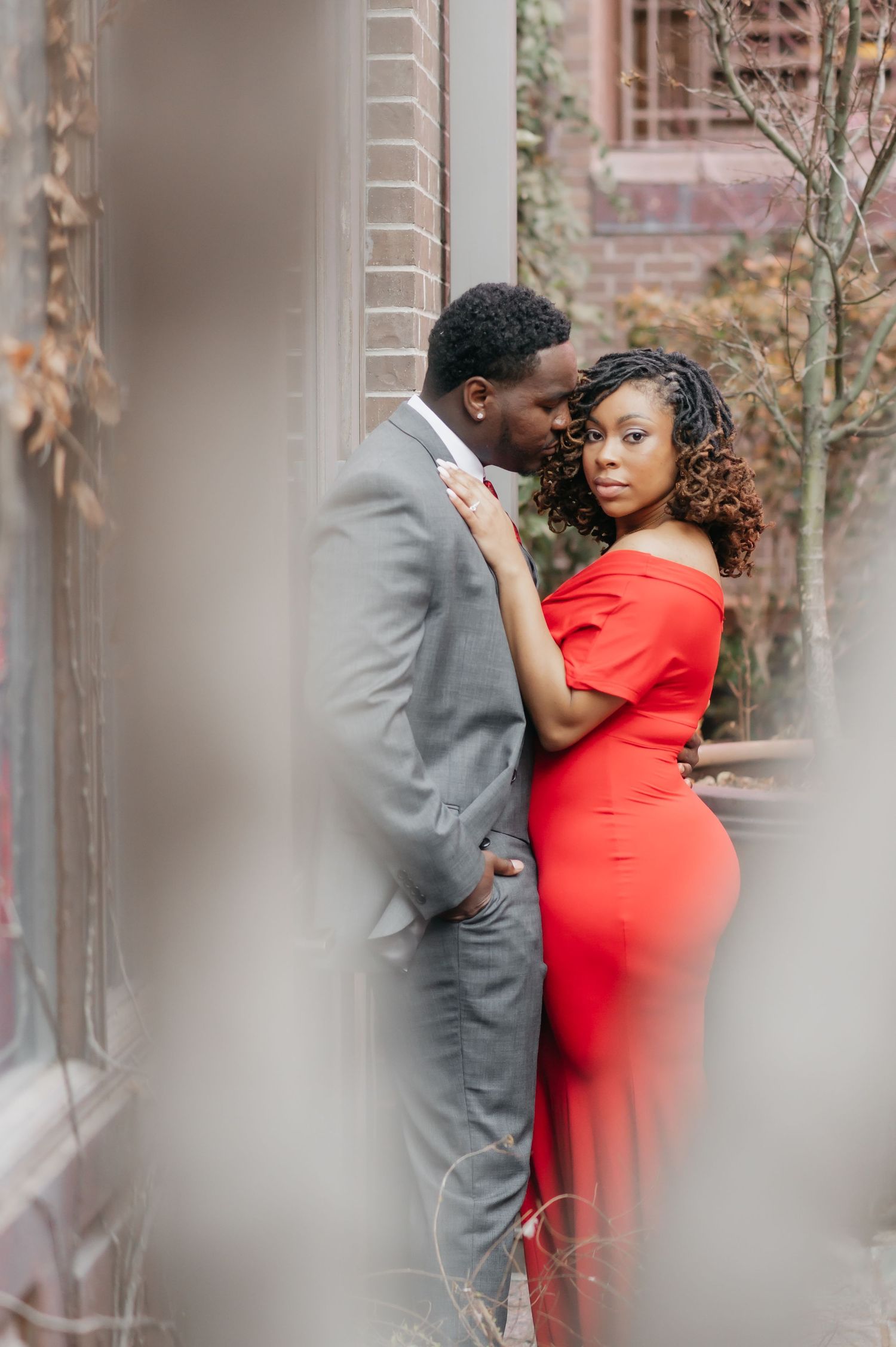 Engaged couple in red dress and gray suit share intimate glance during outdoor engagement photo session.