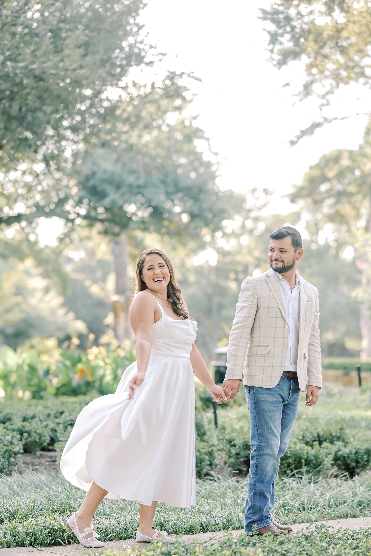 Couple walks hand in hand along a garden path, the woman laughing in a flowing white dress and the man in jeans and a plaid blazer.
