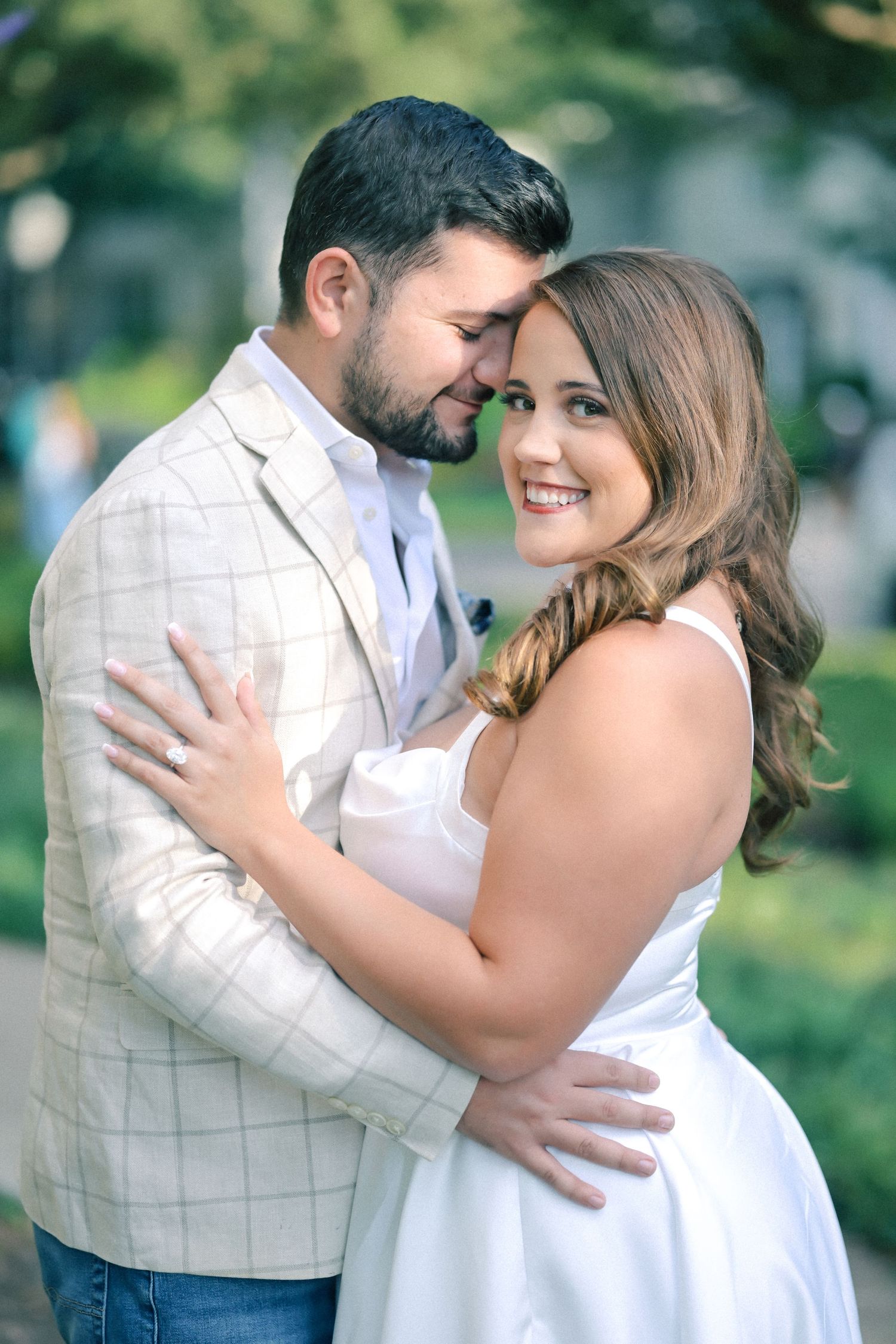 Smiling couple embracing outdoors in a garden, the woman in a white dress and the man in a plaid blazer touching foreheads.