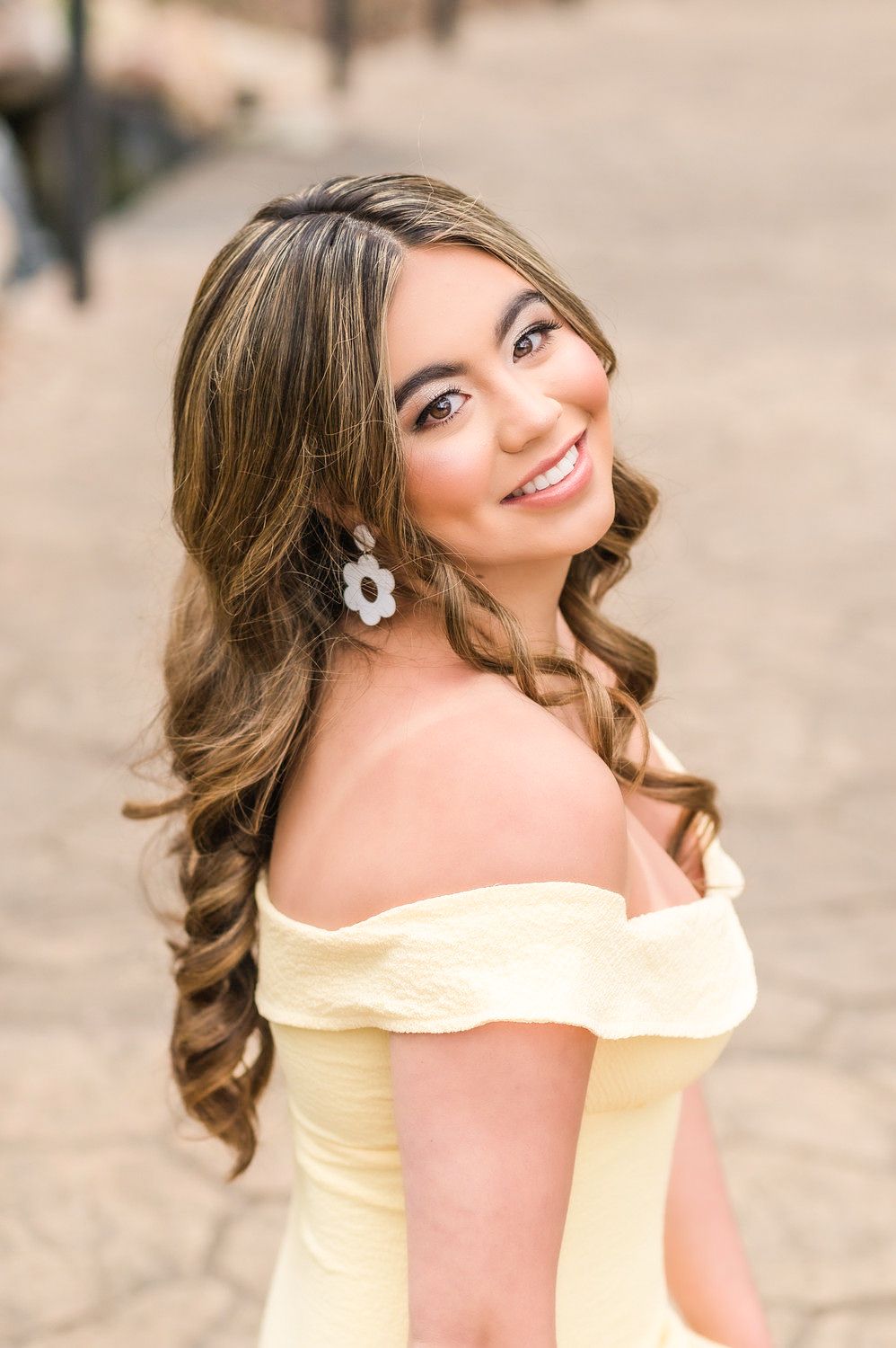 Young woman in yellow off-shoulder dress with floral earrings smiling outdoors.