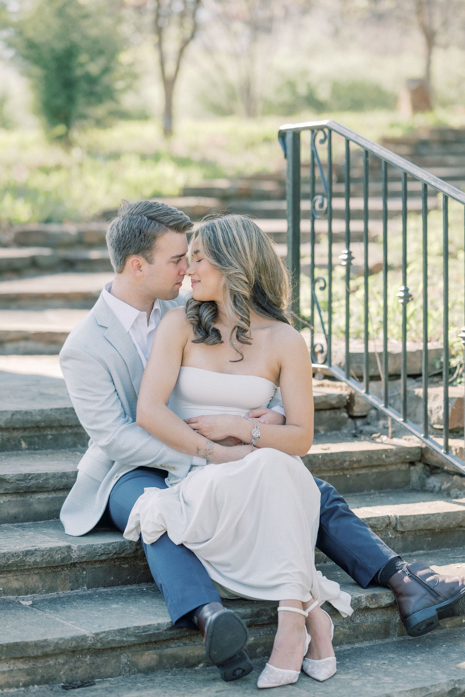 A romantic couple in white clothing shares tender moments while sitting on stone steps in a garden setting.