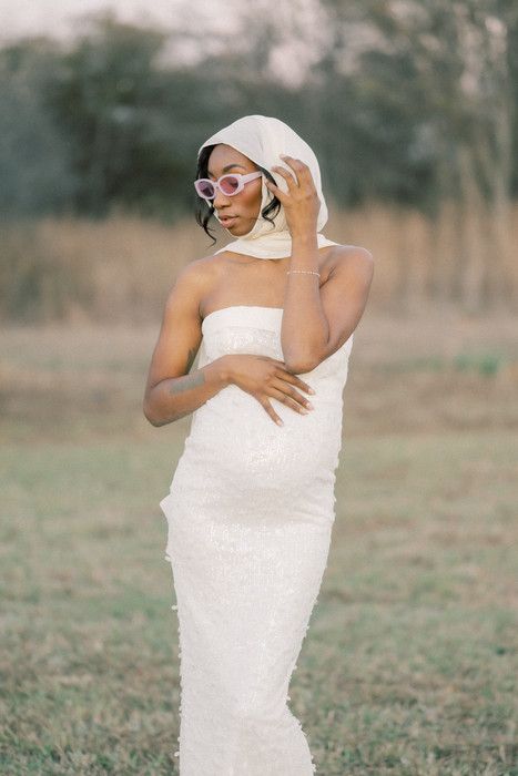 Stylish maternity portrait featuring a woman in white dress and headscarf posing with sunglasses in a natural setting.