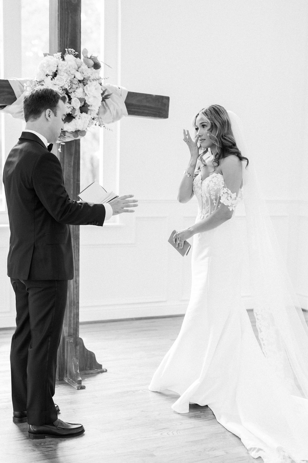 Black and white photo of a bride wiping tears as the groom reads heartfelt wedding vows at a church altar.