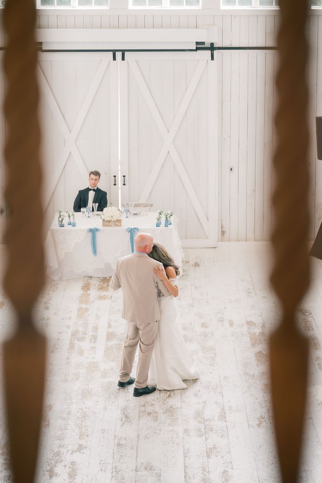 Bride and father share first dance in rustic white barn wedding venue with groom seated at sweetheart table.