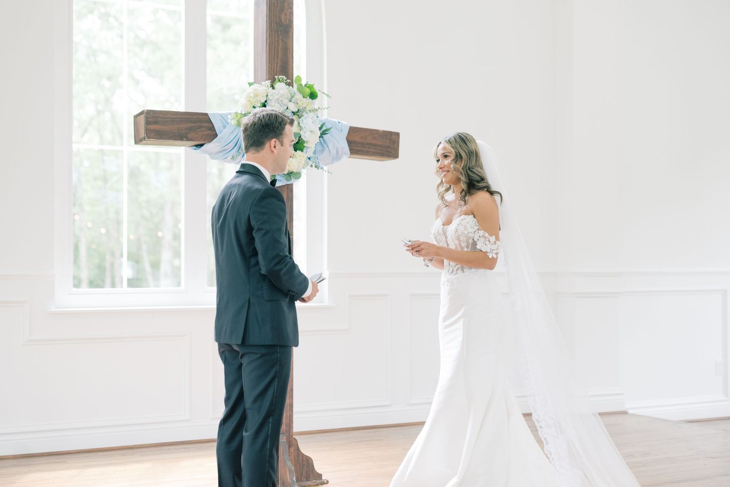 Bride and groom exchange vows before a wooden cross in a bright white chapel wedding ceremony.