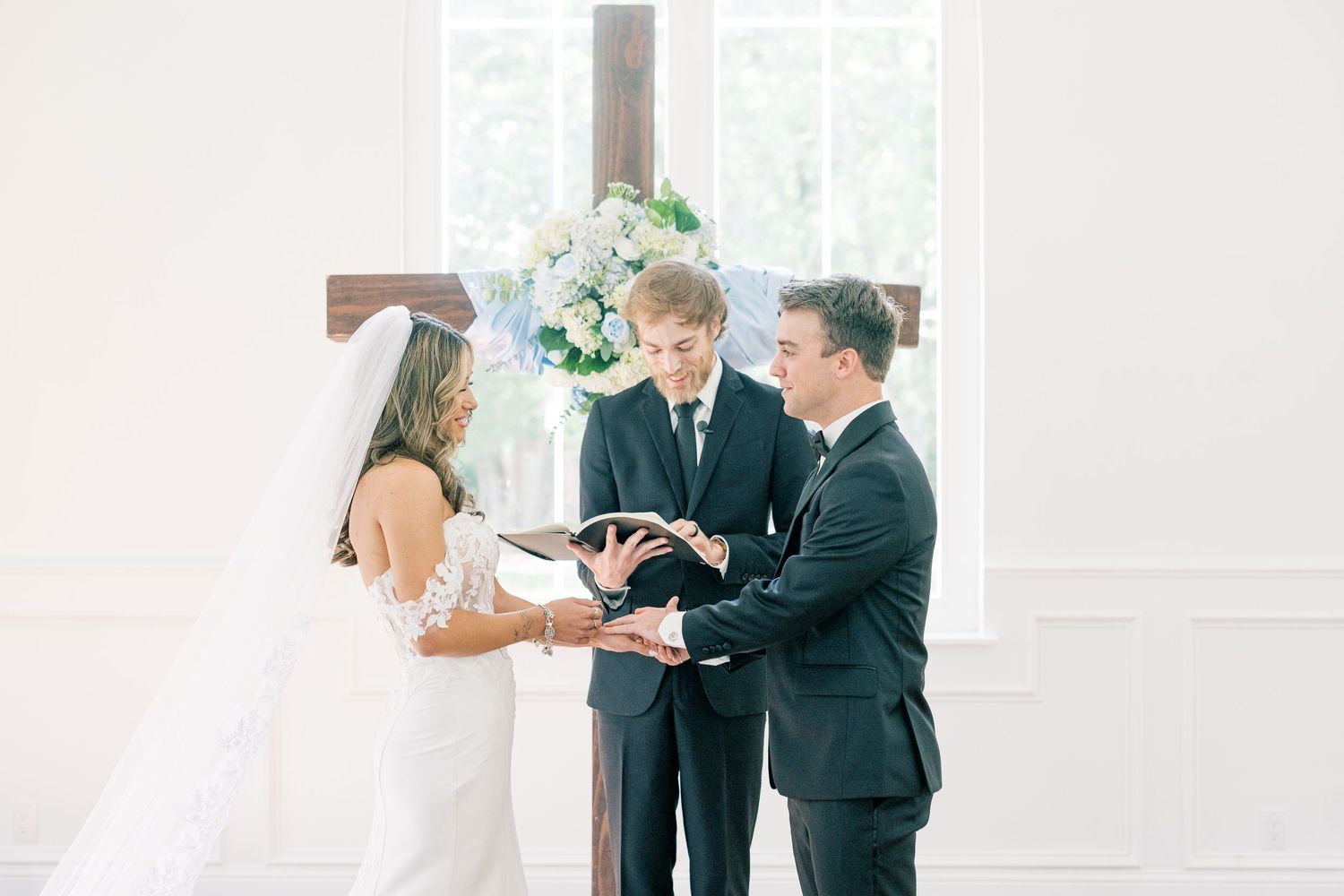 Bride and groom exchanging vows with officiant at white chapel altar with floral arrangements.