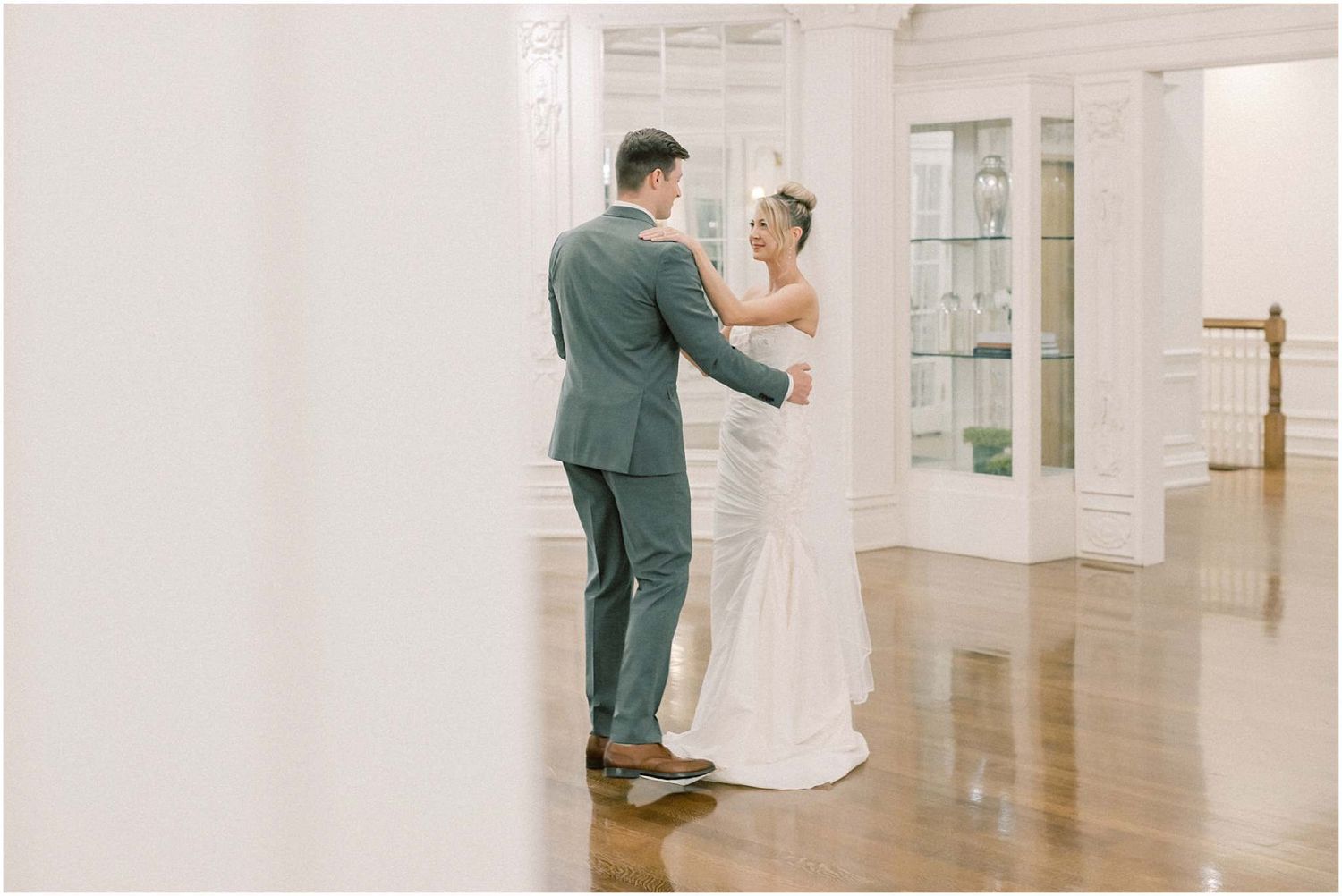 Intimate wedding dance moment captured in bright white ballroom with hardwood flooring.