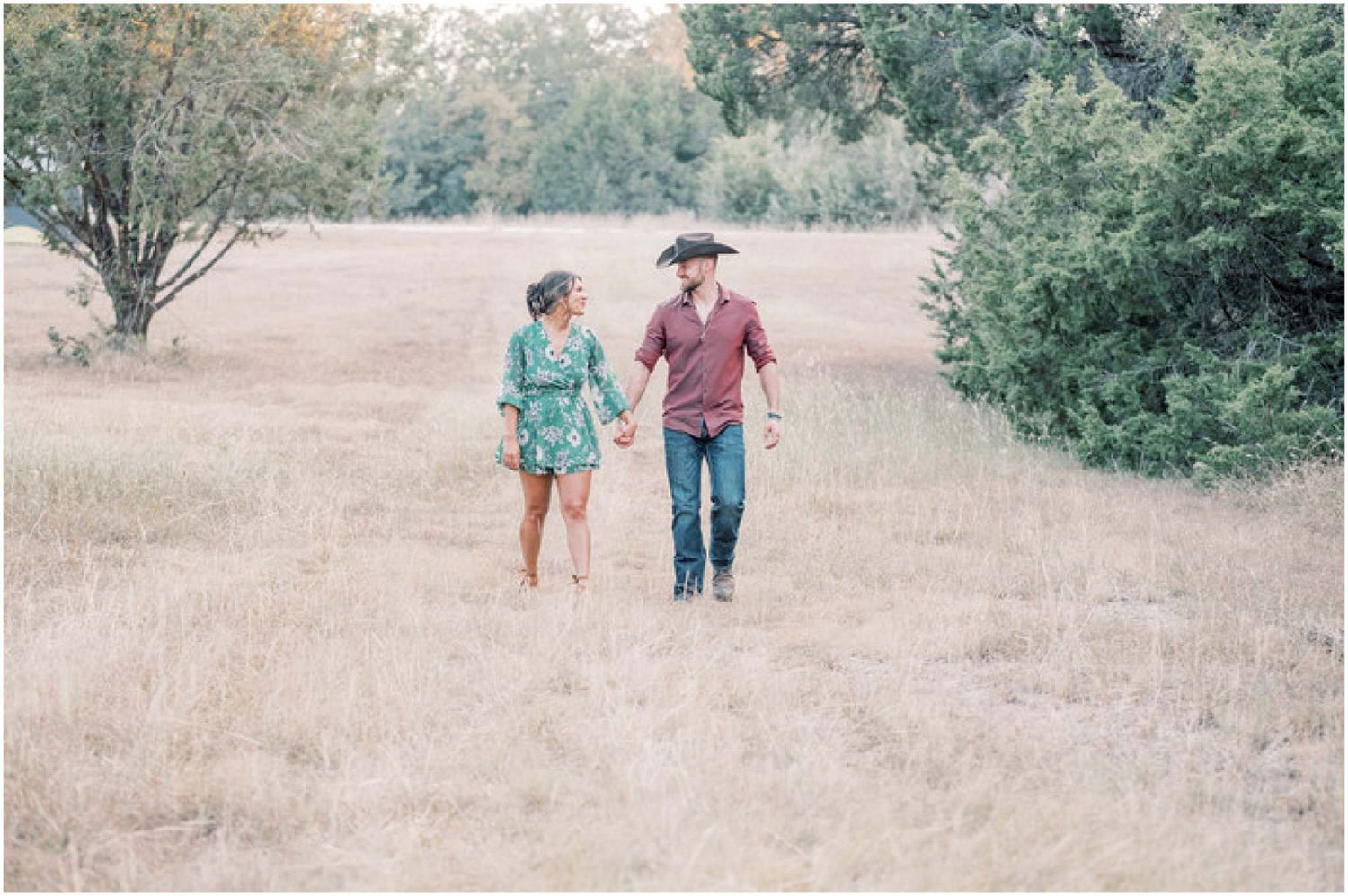 A couple walks together in a grassy field on a sunny day, wearing casual attire.