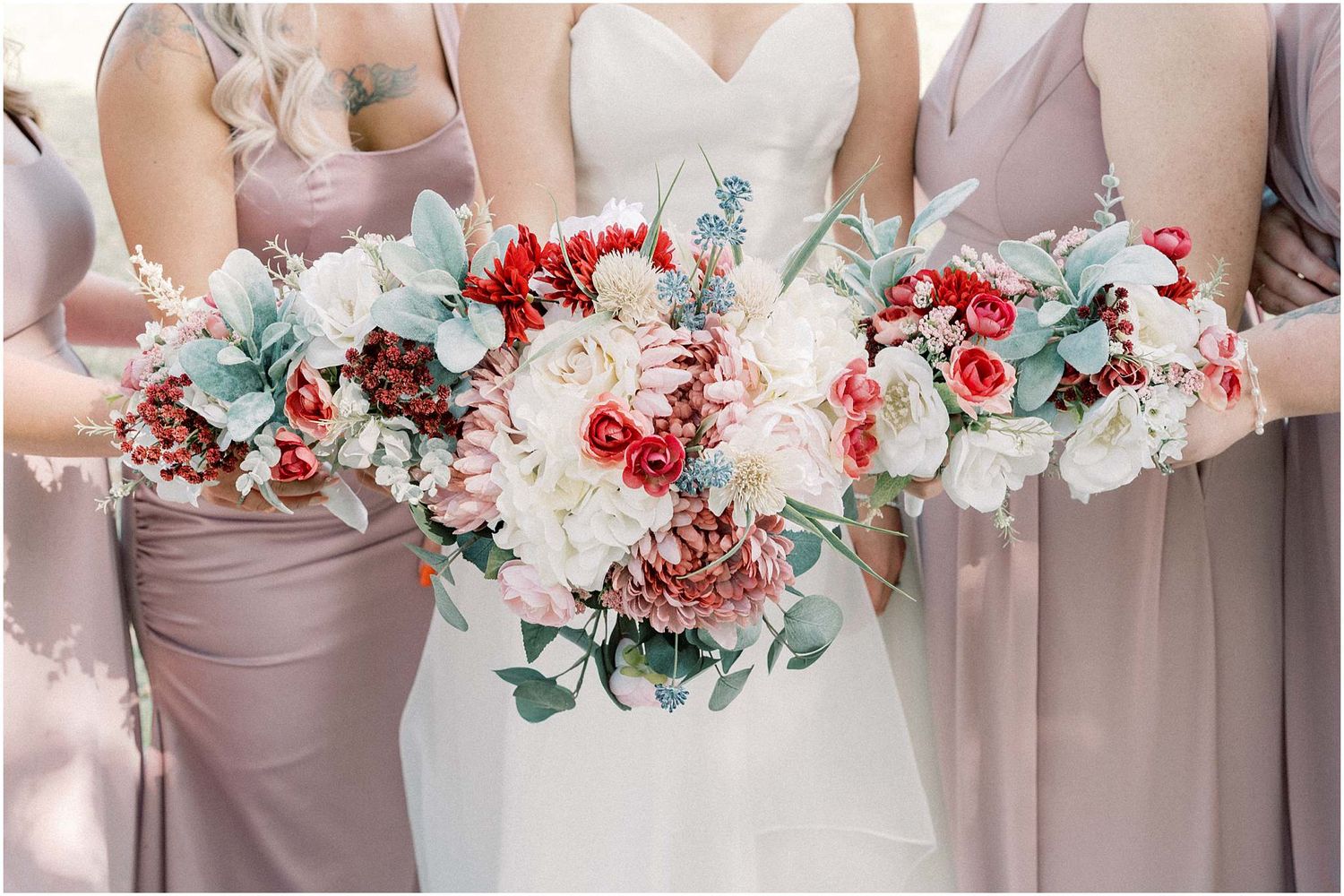 Bridesmaids in dusty rose dresses hold elegant bouquets with red and white flowers against a light background.