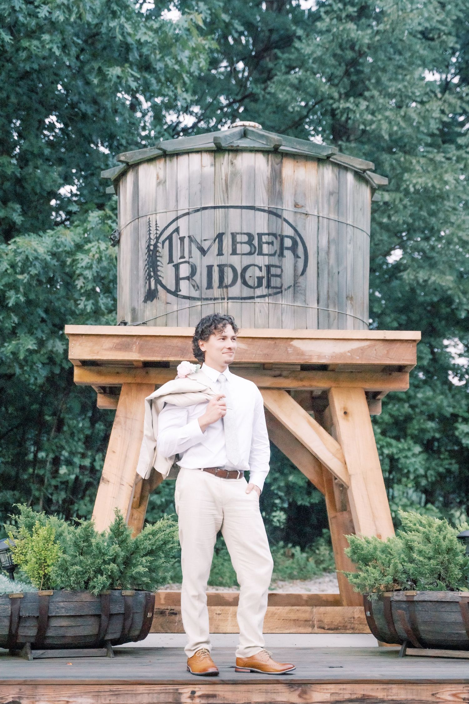 Man in white outfit standing before a rustic wooden Timber Ridge water tower surrounded by lush greenery.
