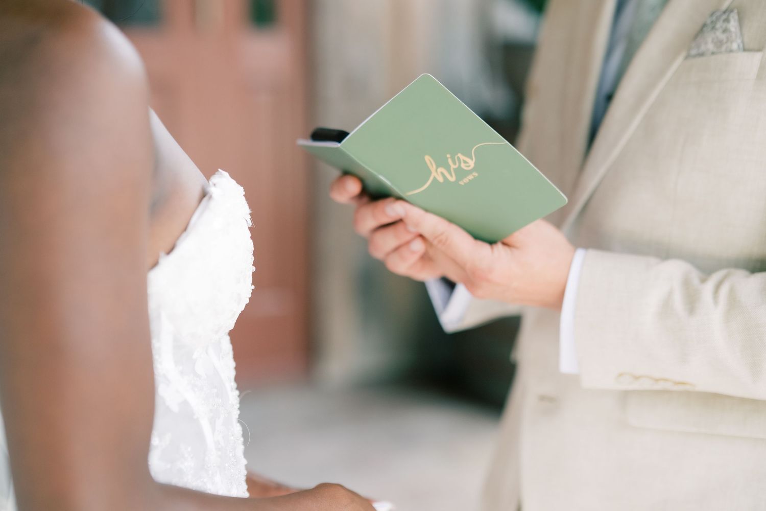 Groom in beige suit reads vows from a sage green booklet during an outdoor wedding ceremony.