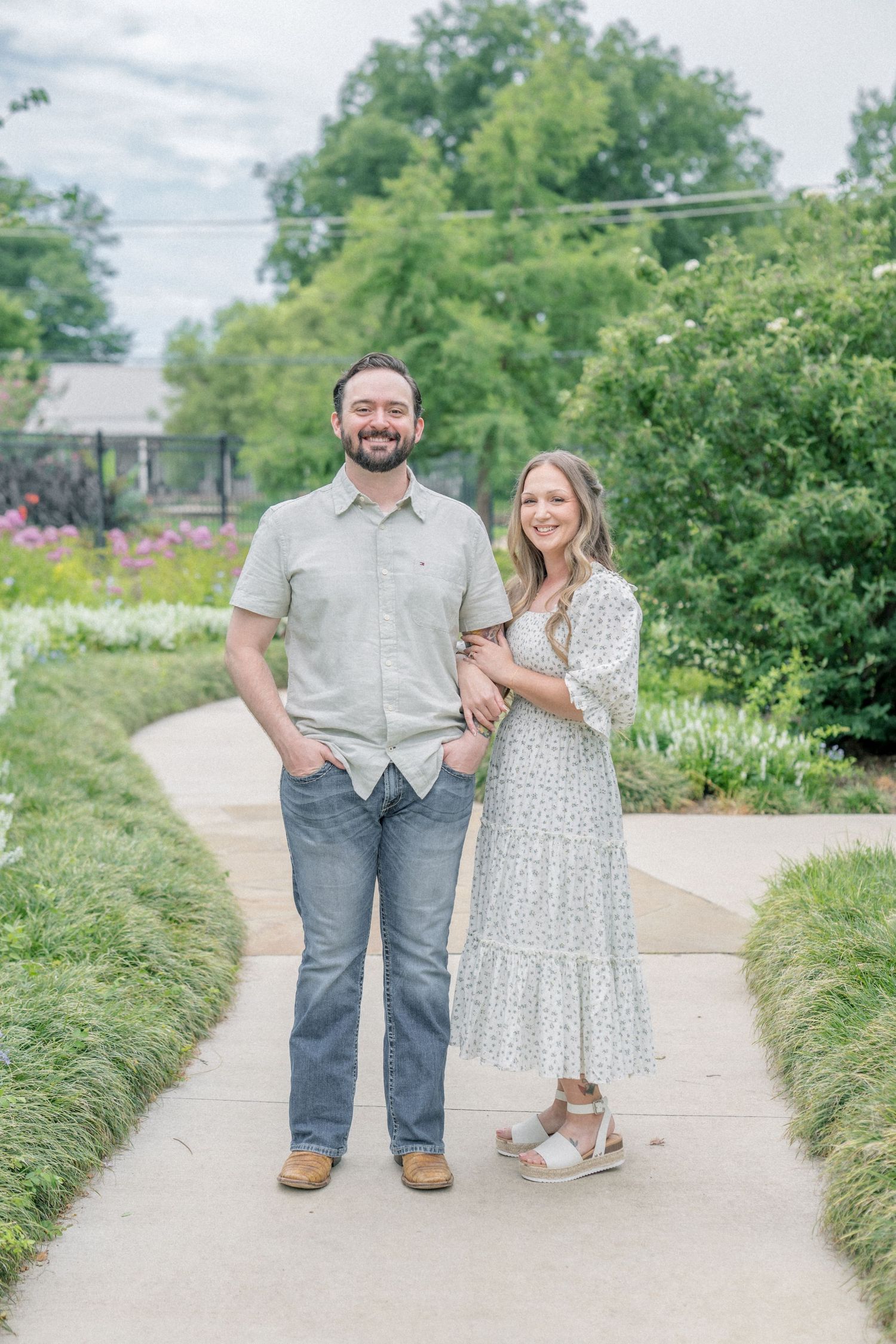 A smiling couple poses on a garden path, man in gray shirt and jeans, woman in white floral midi dress.