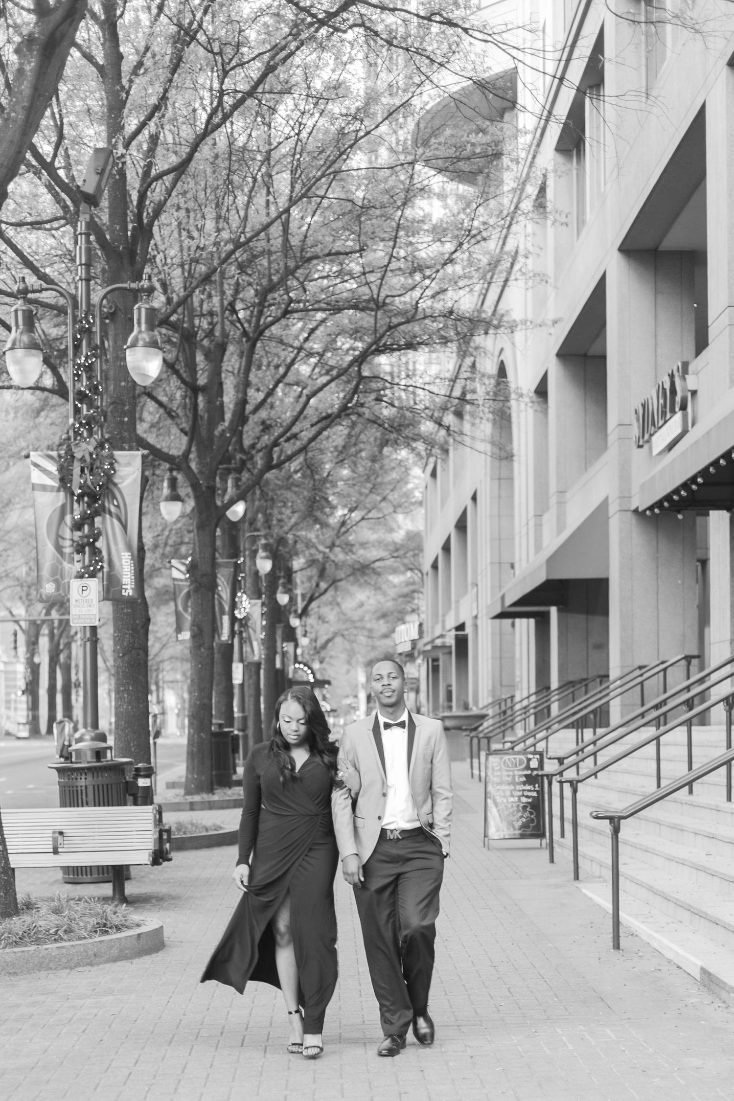 Elegant Black couple walking hand-in-hand on urban sidewalk, woman in black gown and man in suit, black and white photo.
