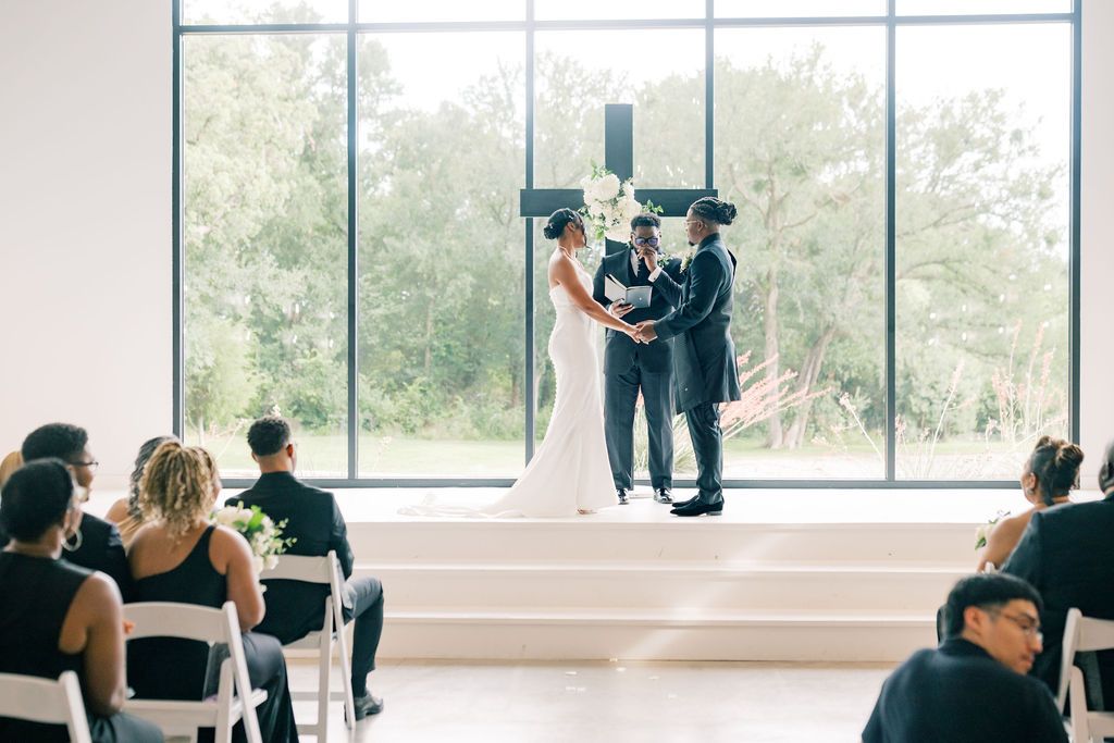 Couple exchanges vows at modern wedding ceremony with large windows, cross backdrop, and seated guests in white chairs.