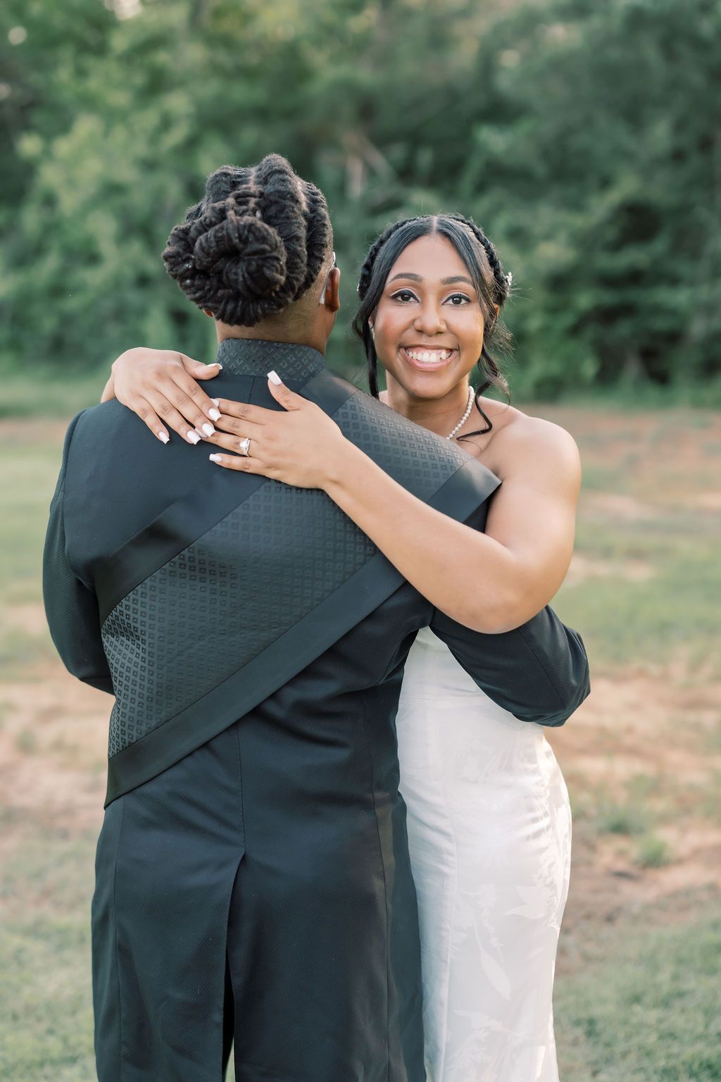 A smiling bride in a white dress embraces her partner in a black suit outdoors during a wedding celebration.