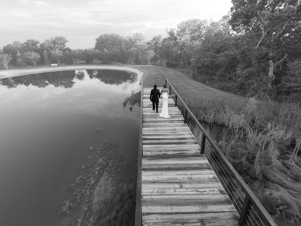 Black and white photo of a couple walking on a wooden boardwalk beside a calm lake surrounded by lush trees.