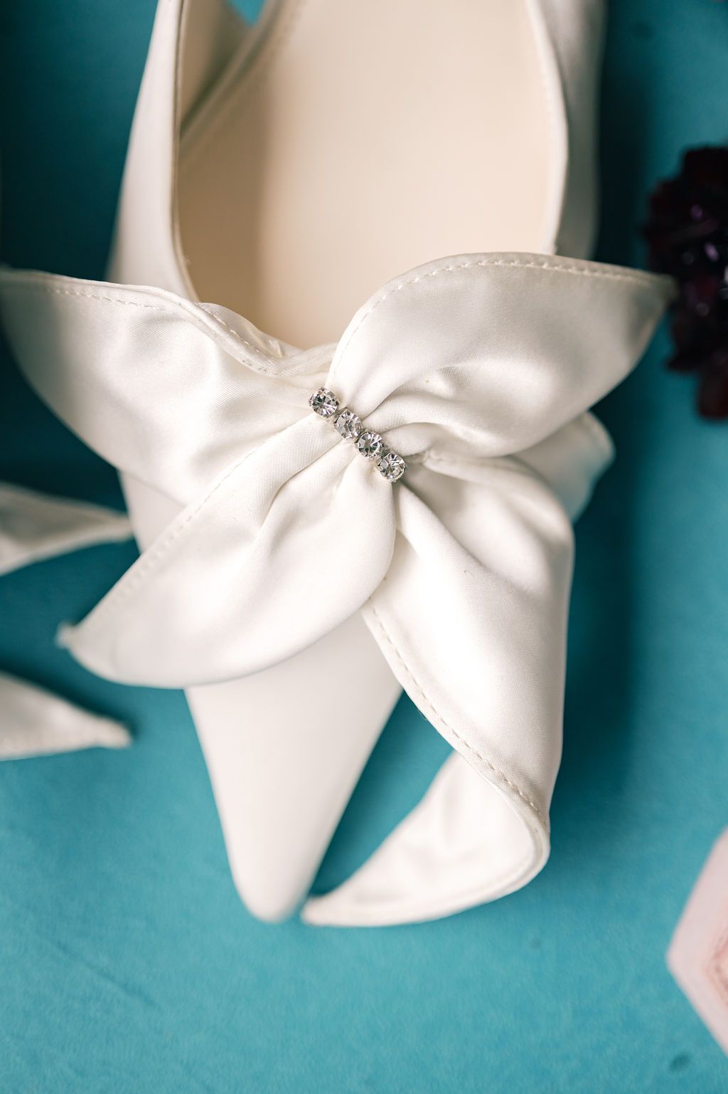 Elegant white satin bridal heels with large bow and crystal rhinestone embellishment on teal background.