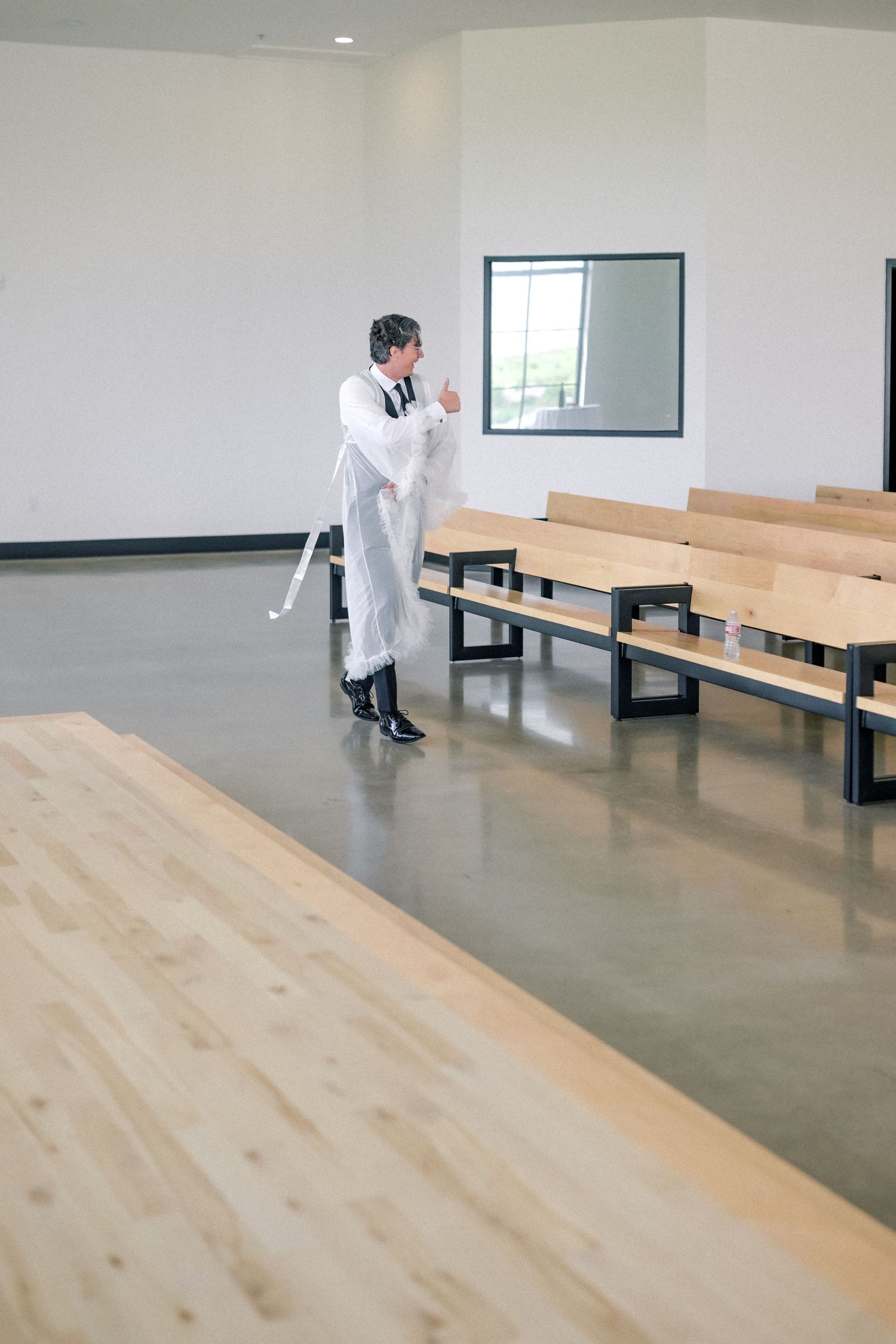 Officiant in white robe reviewing notes among wooden pews in a minimalist modern wedding ceremony space.