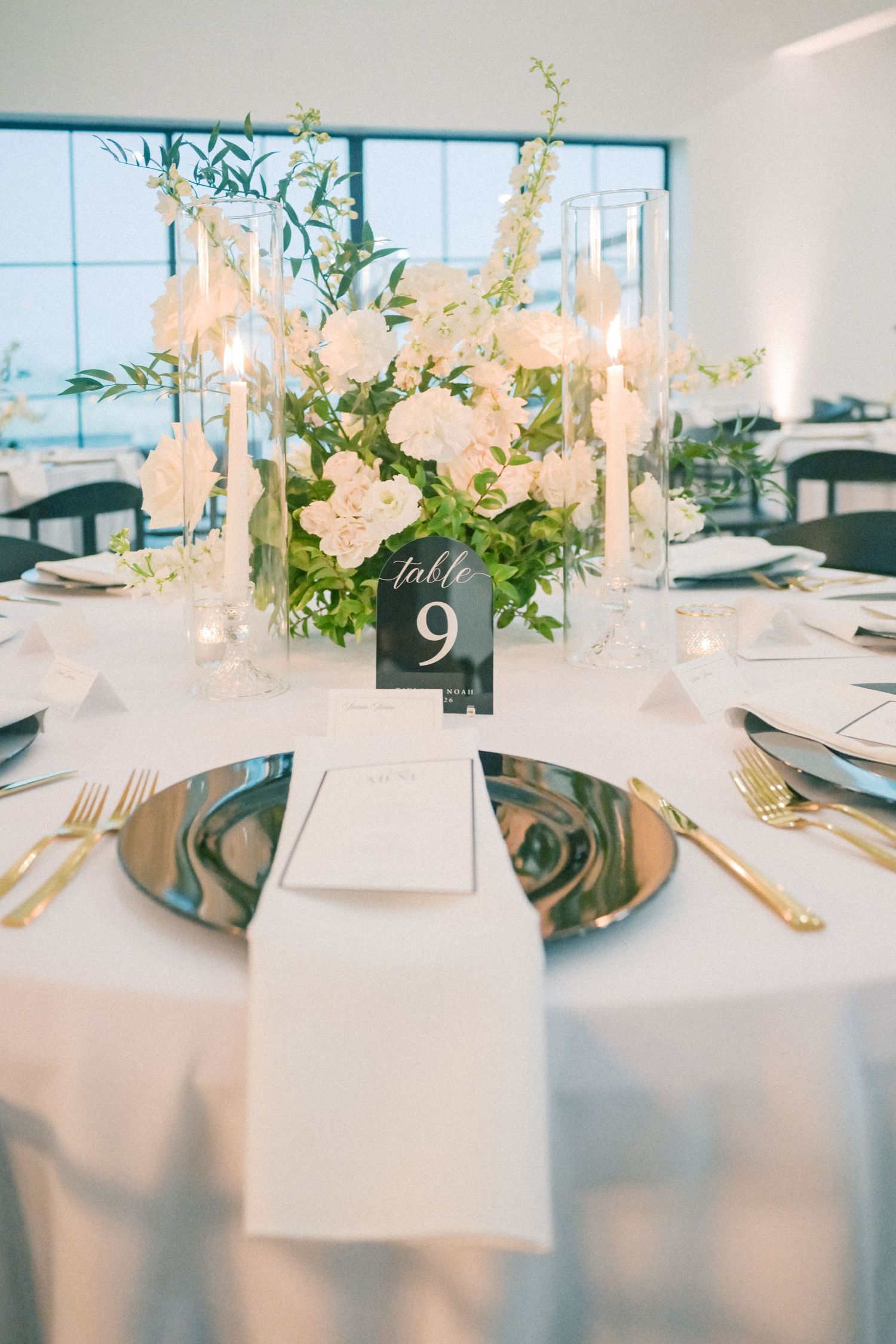 Elegant wedding reception table nine features white floral centerpiece, gold cutlery, and charger plates.