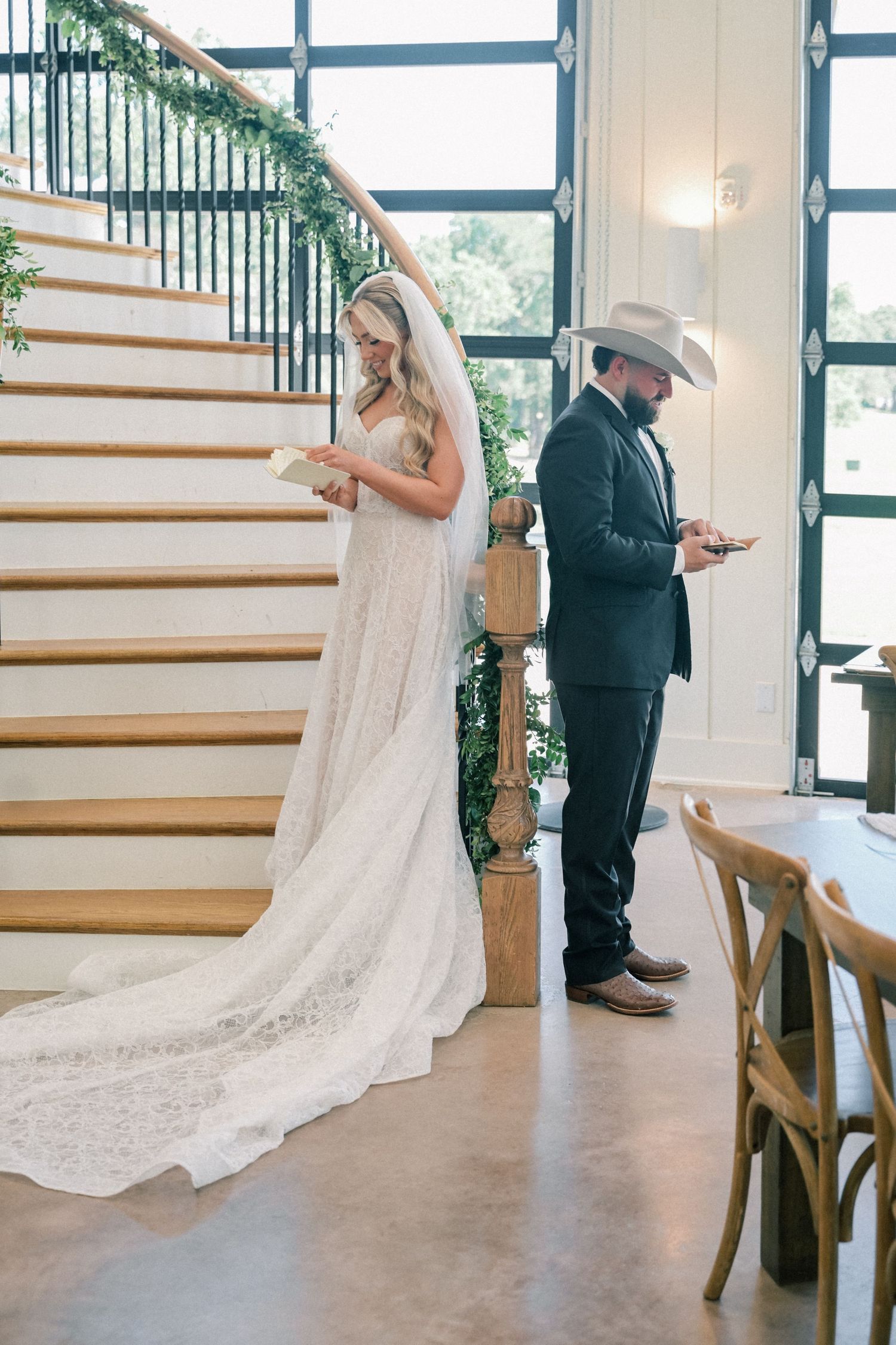 Bride in lace gown and groom in suit with cowboy hat share private letters back-to-back near elegant staircase.