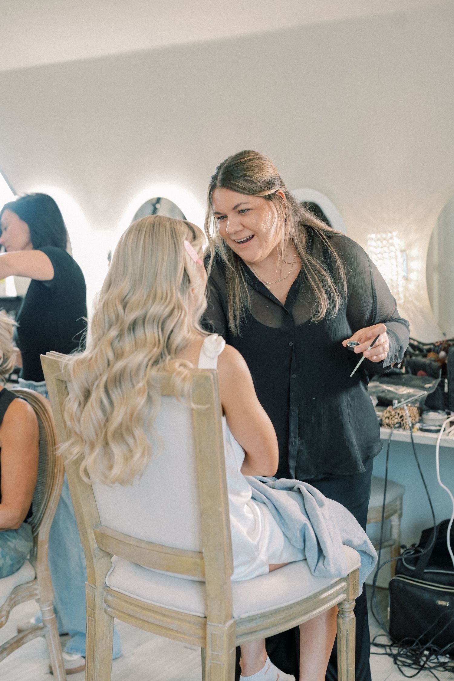 Makeup artist in black top laughing with blonde bride during wedding day preparations indoors.