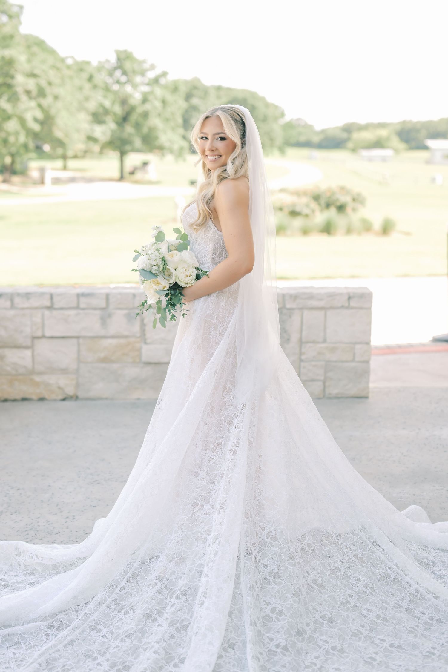 Radiant bride in lace gown and long veil holding white floral bouquet outdoors on sunny day.