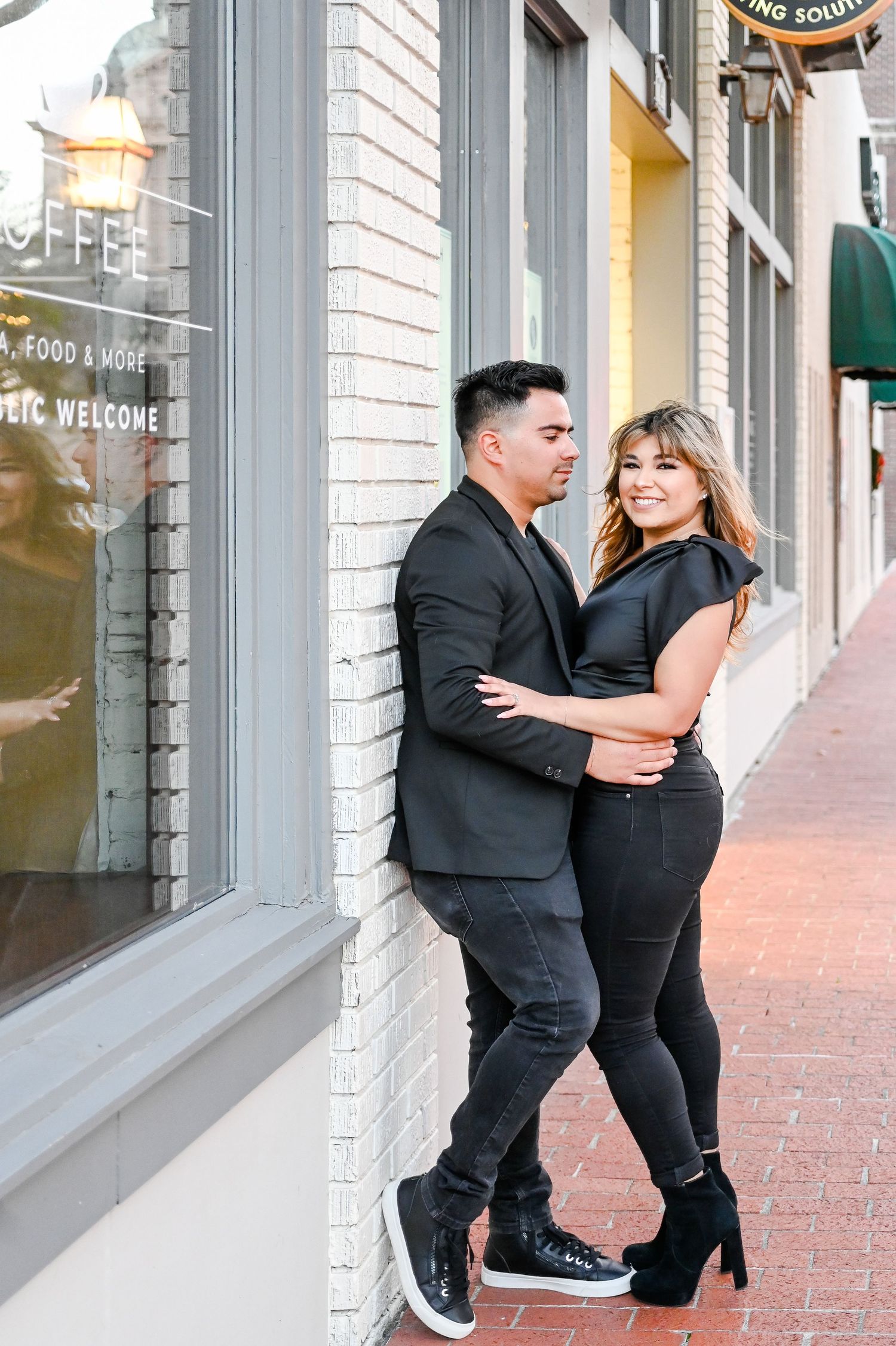 Couple in matching black outfits posing against a brick wall on a city sidewalk, sharing a romantic moment.