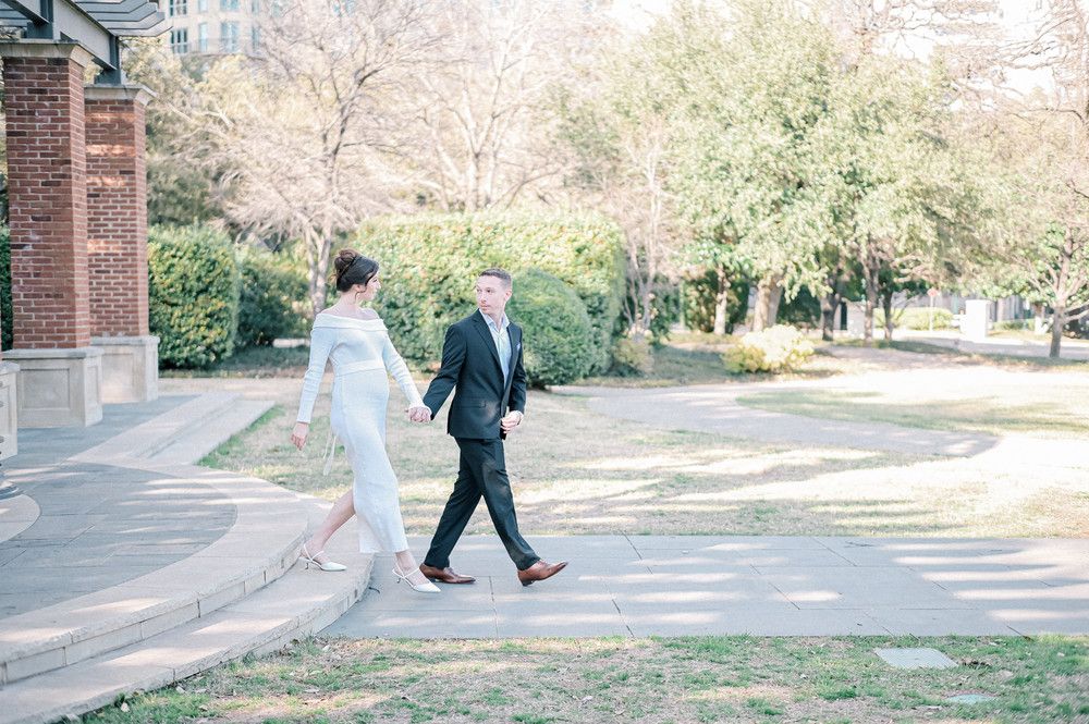 Bride and groom walking hand in hand along a sunlit park pathway, bride in white gown, groom in dark suit.