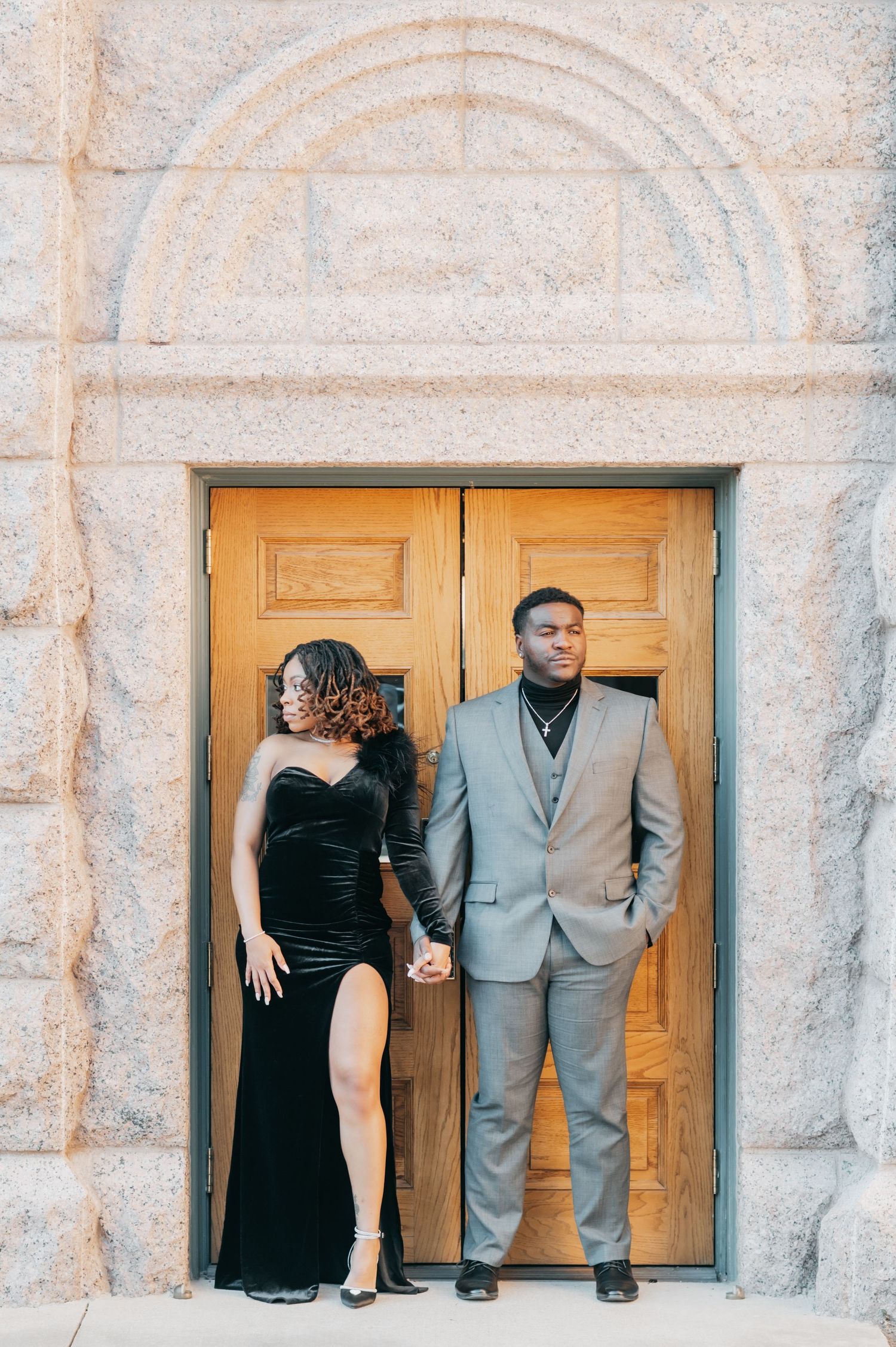 Stylish couple holding hands before ornate wooden doors, she wears a black velvet dress, he wears a gray suit.