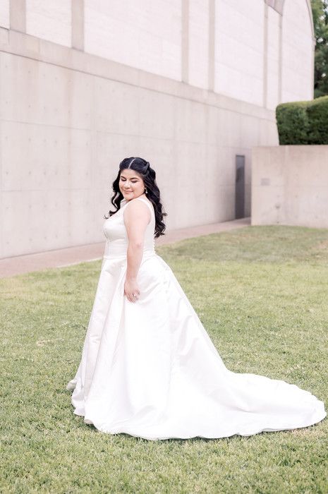 Bride in a white ball gown with a long train poses gracefully on green grass beside a modern building.