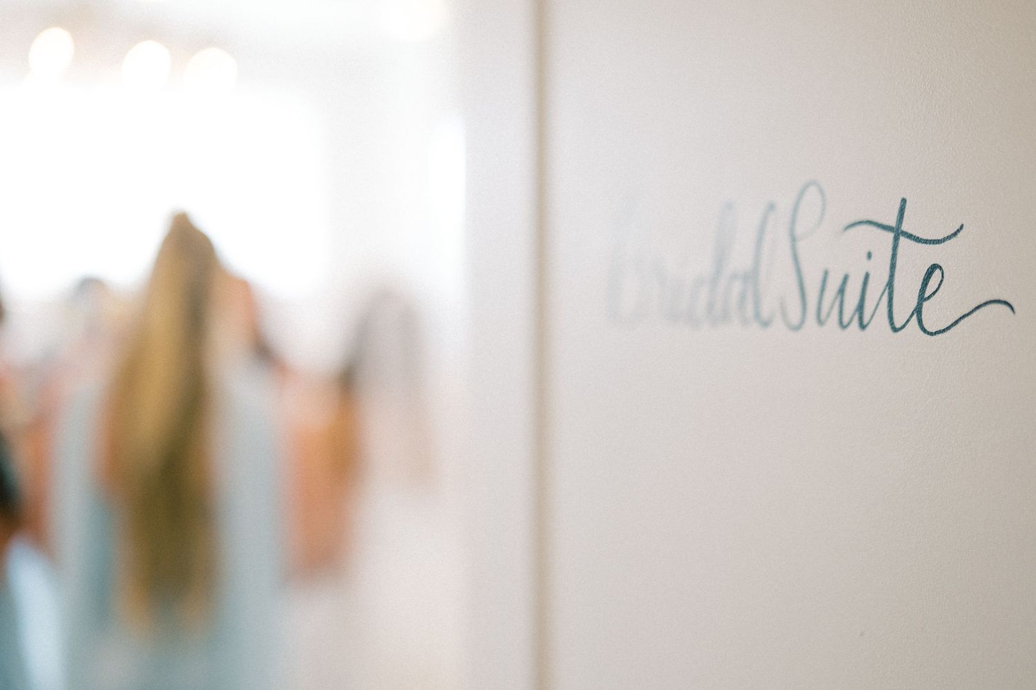 Elegant bridal suite sign in script lettering on a white wall with blurred bridesmaids in background.