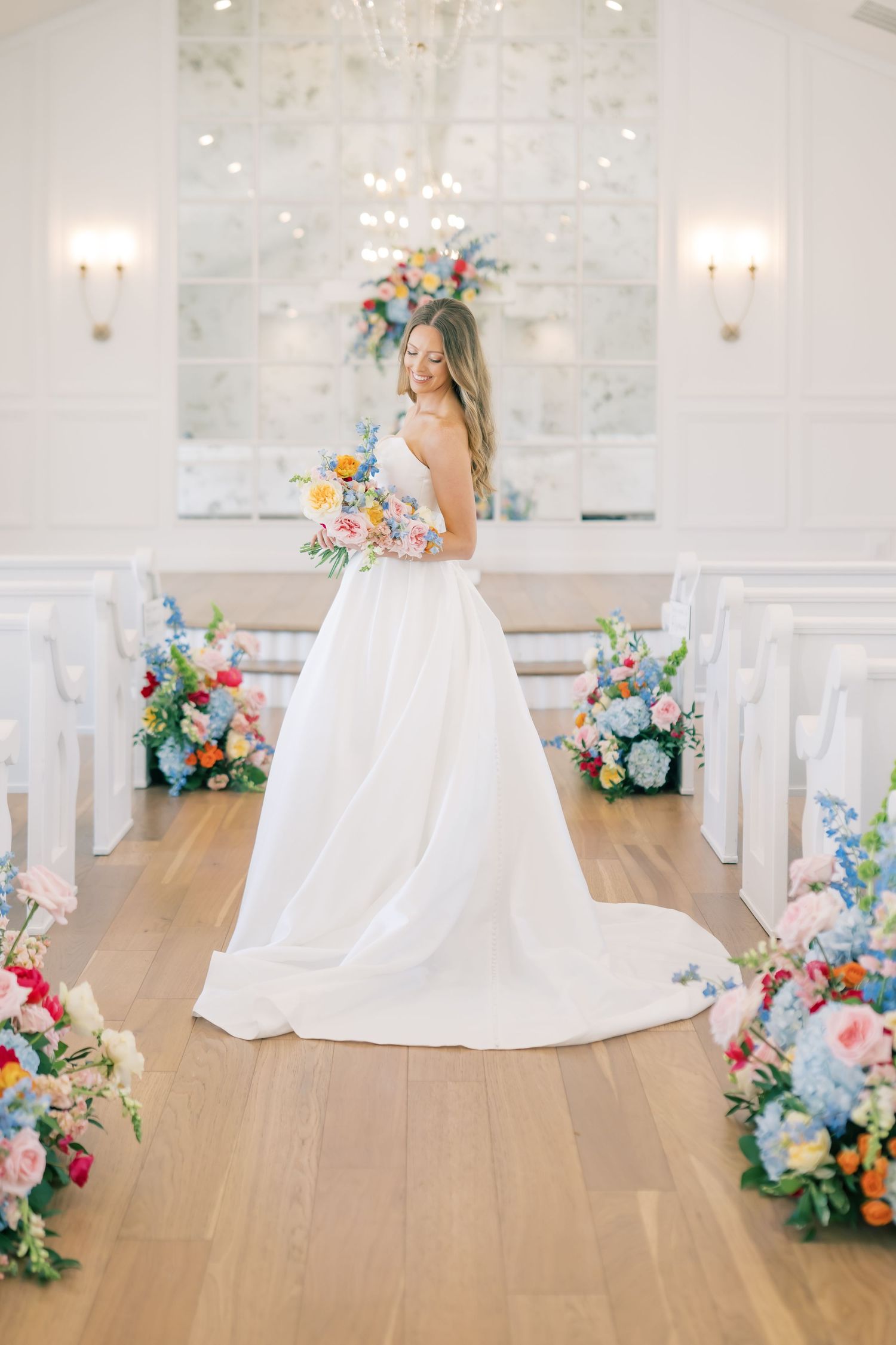 Bride in white ball gown holding colorful bouquet stands in aisle of chapel decorated with vibrant floral arrangements.
