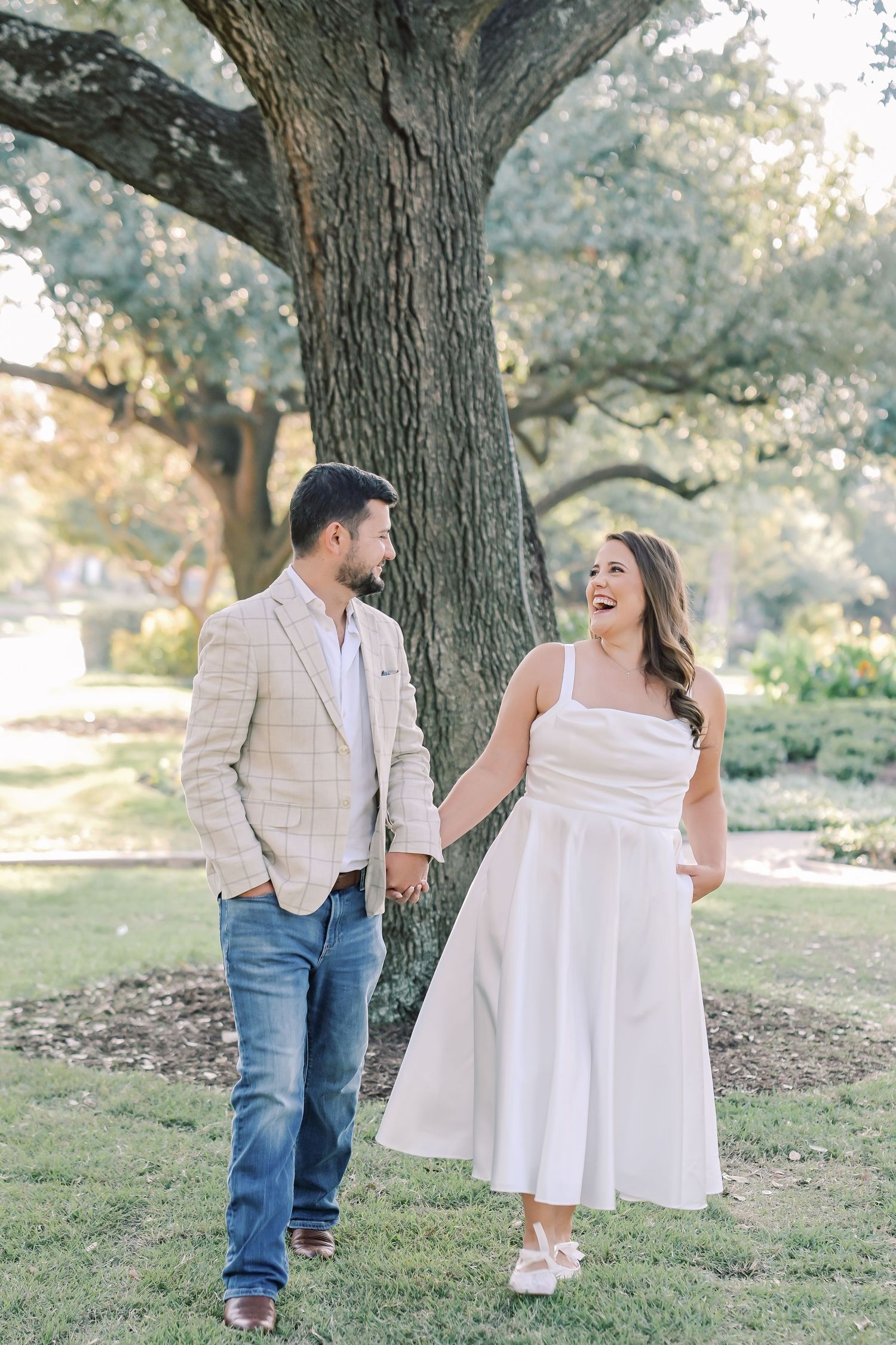 Laughing couple holding hands and walking toward the camera beneath a large oak tree in a green park for an engagement session.