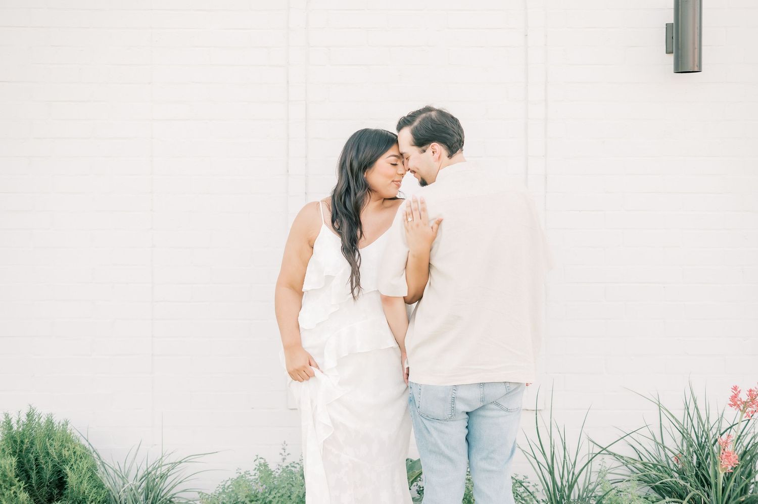 A couple embraces in a romantic outdoor photoshoot with lush greenery and white outfits.