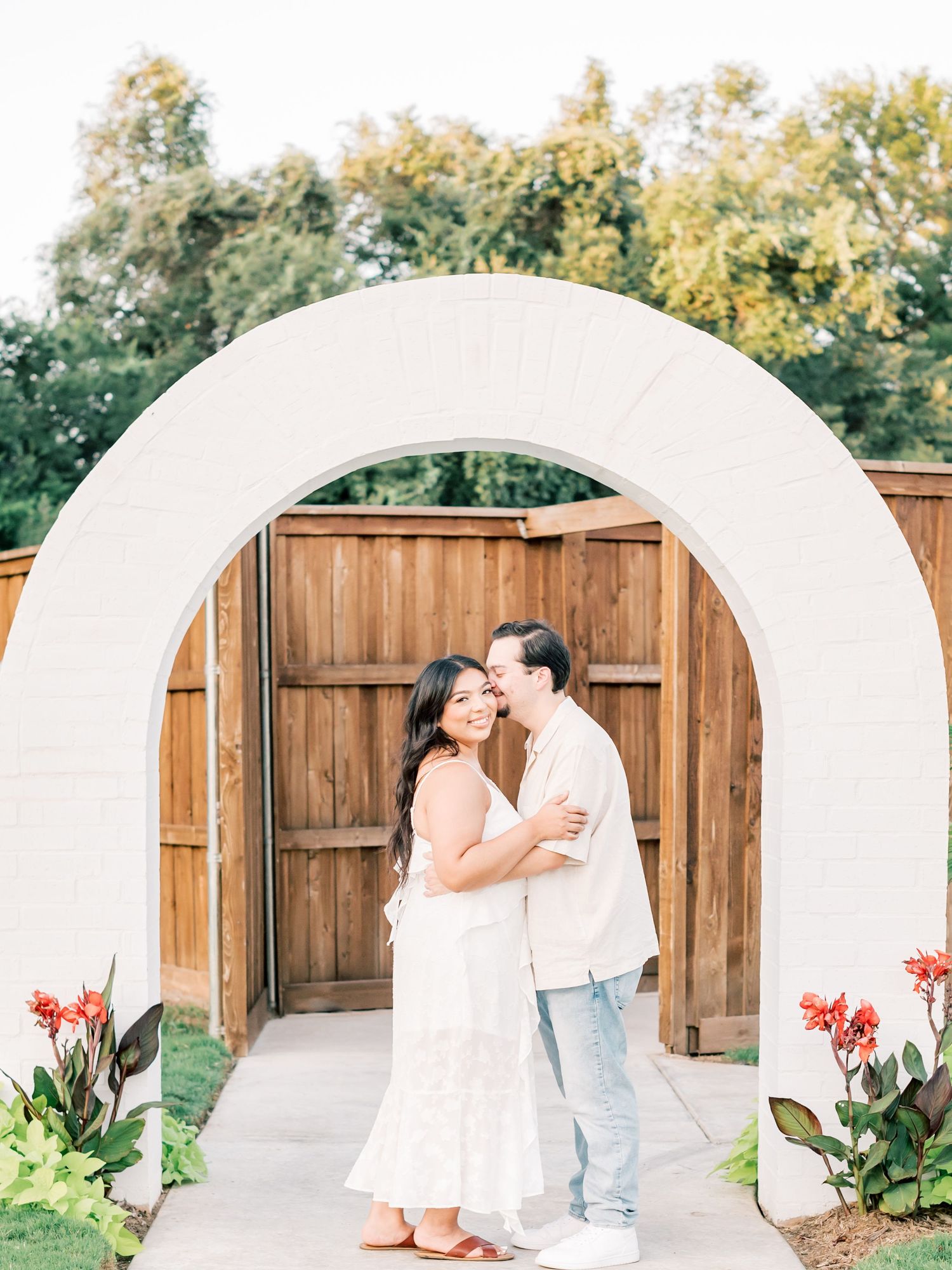 A series of five romantic photos showing a couple in white outfits embracing in front of white arched doorways in a garden setting.
