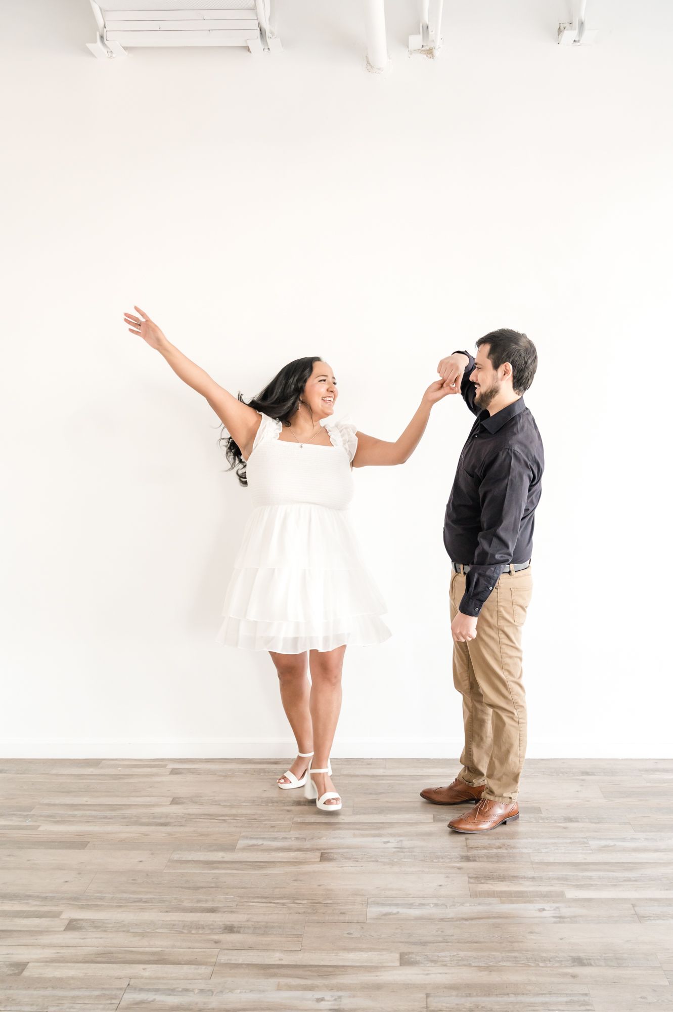 A playful couple dances and poses in a bright white studio with wooden floors.