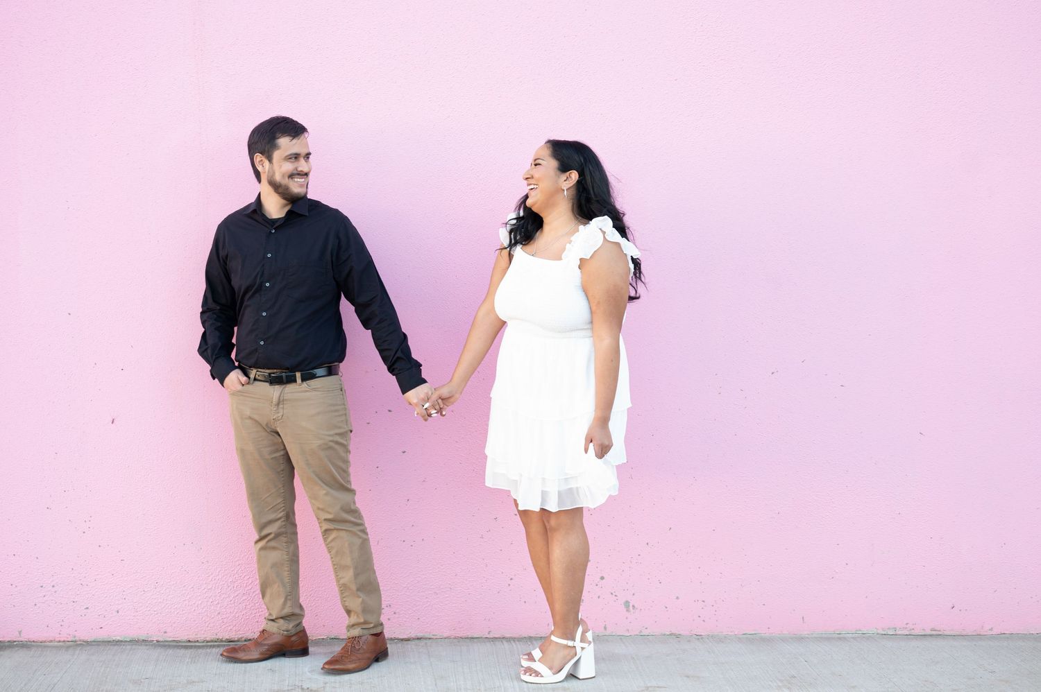 A couple holding hands and laughing against a pink wall, with her in a white dress and him in a dark shirt.