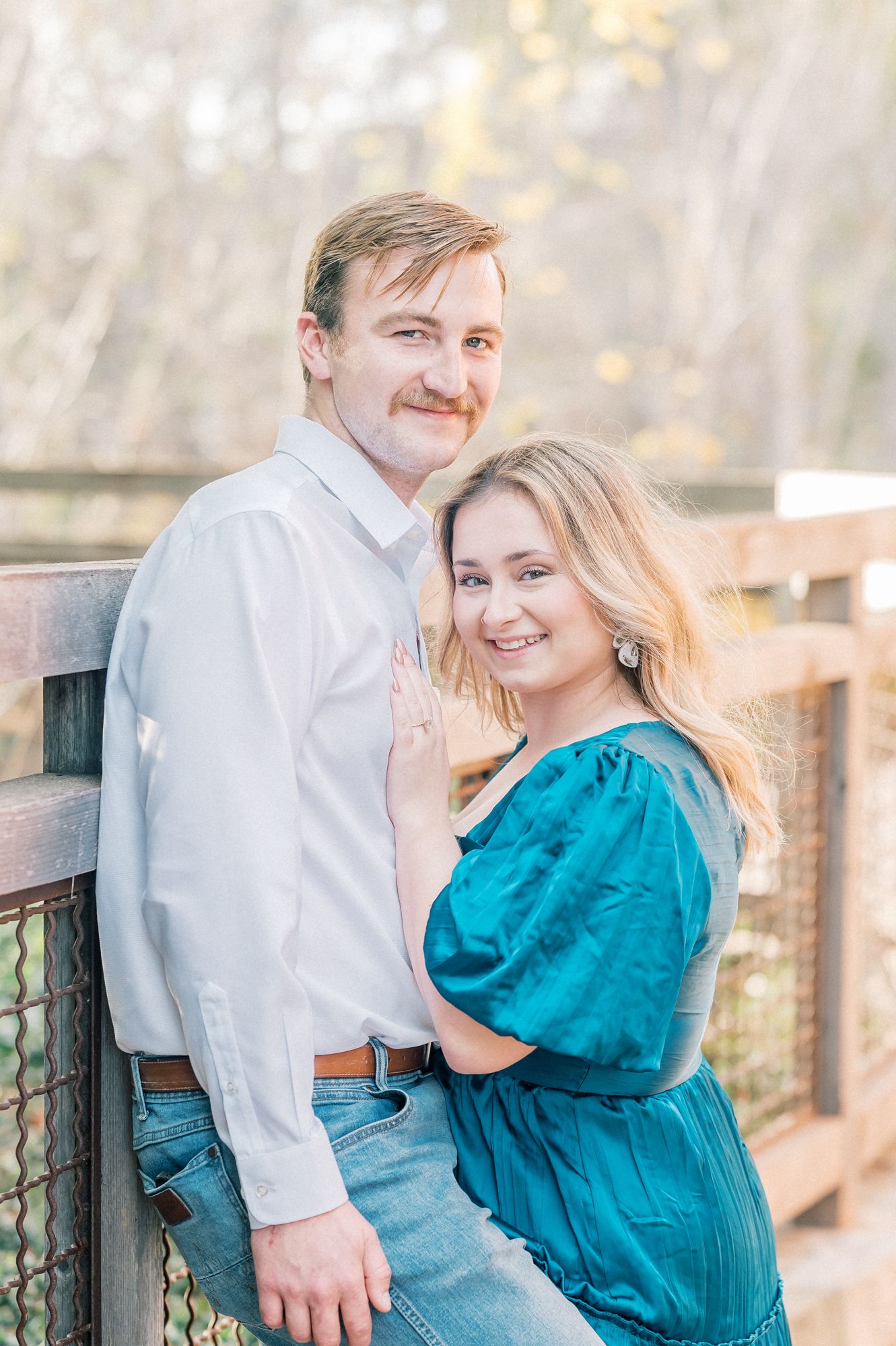 A romantic engagement photo of a couple in casual attire posing together on a wooden deck outdoors.