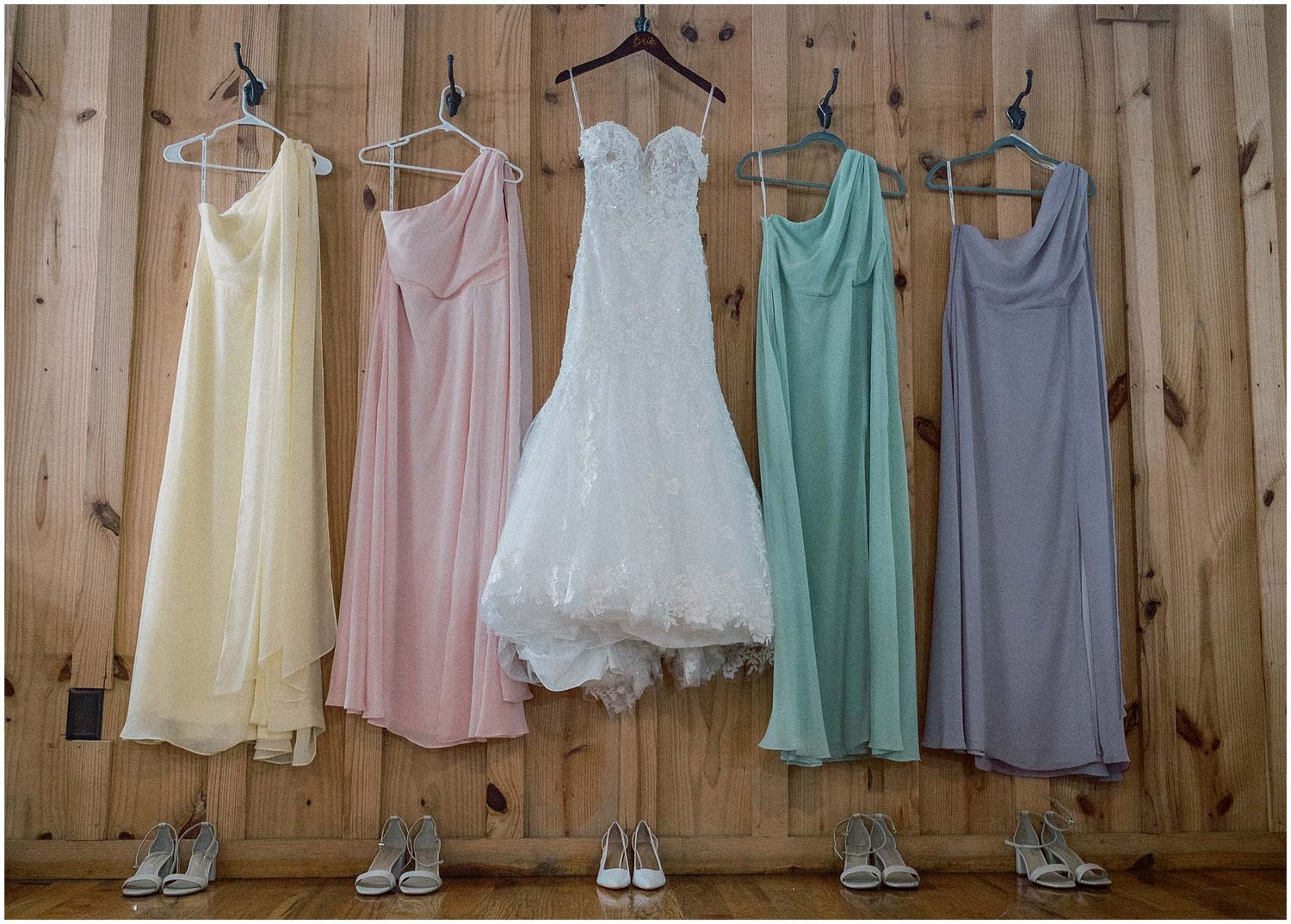 Bridesmaid dresses in pastel colors hanging on wooden wall display with matching shoes below.