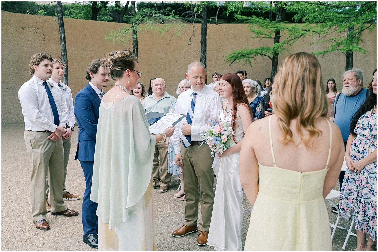 Wedding guests gather outdoors under trees near a beige wall during a summer celebration.