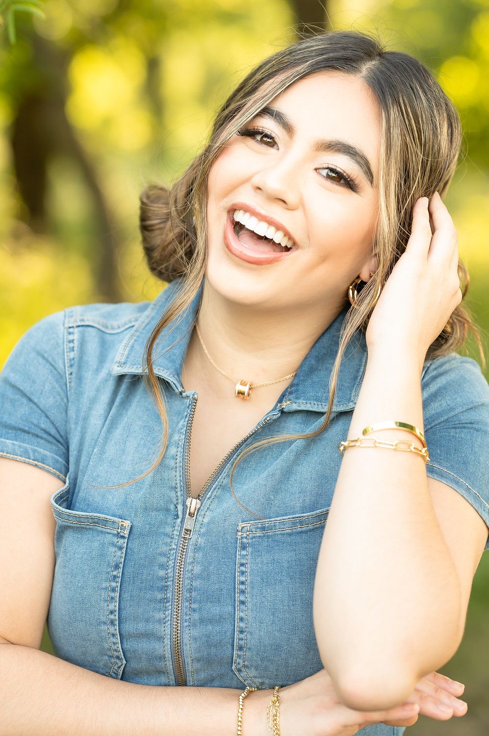 Young woman laughing joyfully in denim dress with gold jewelry outdoors.
