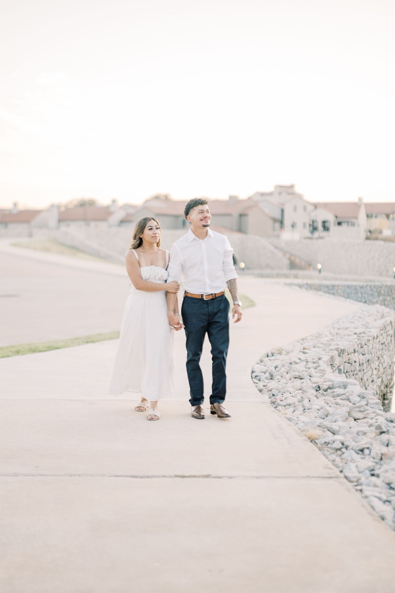 Happy couple walking hand in hand along a stone pathway during a bright outdoor engagement session.
