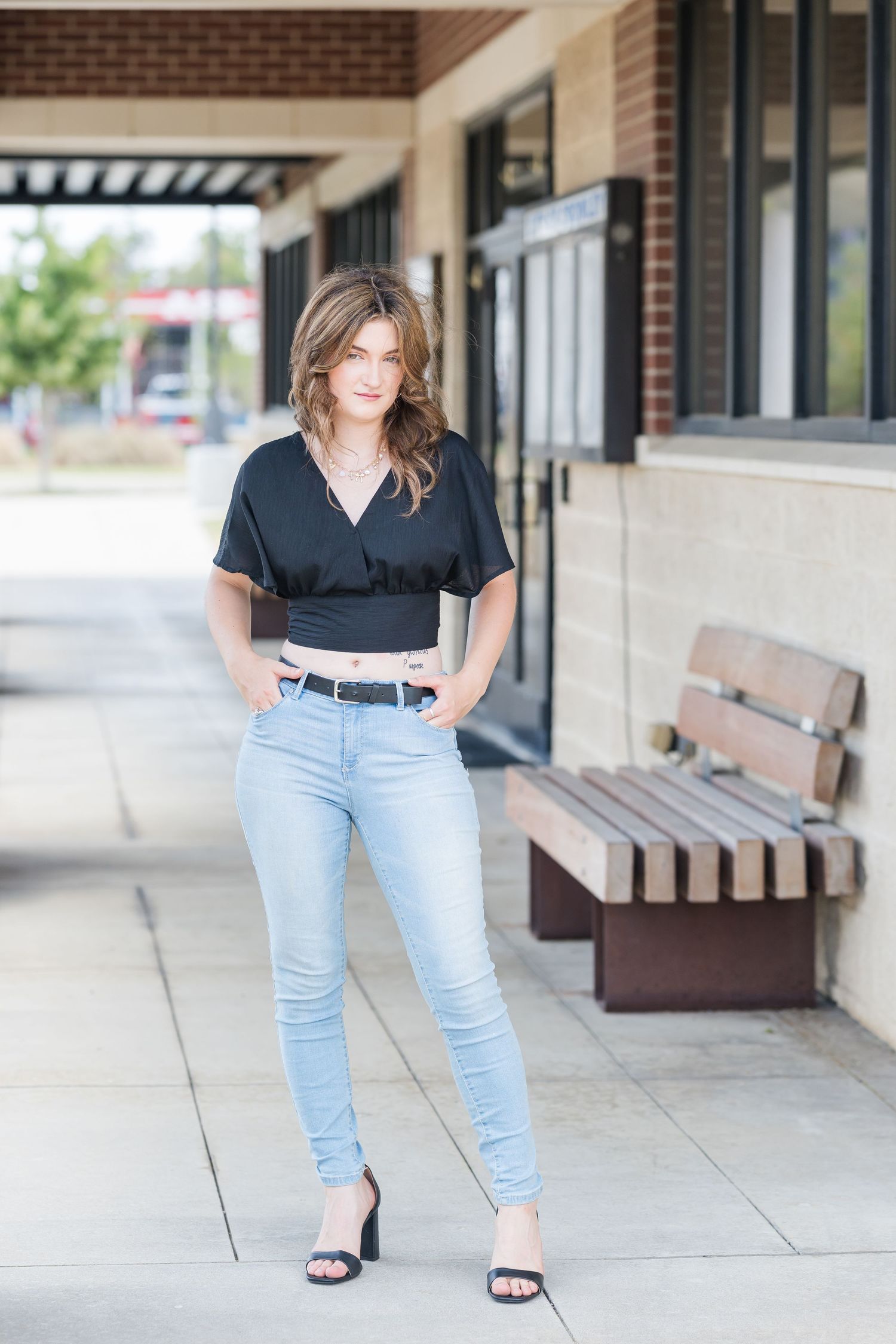 Young woman in black crop top, light jeans, and heels poses on a sidewalk near a brick building and wooden bench.